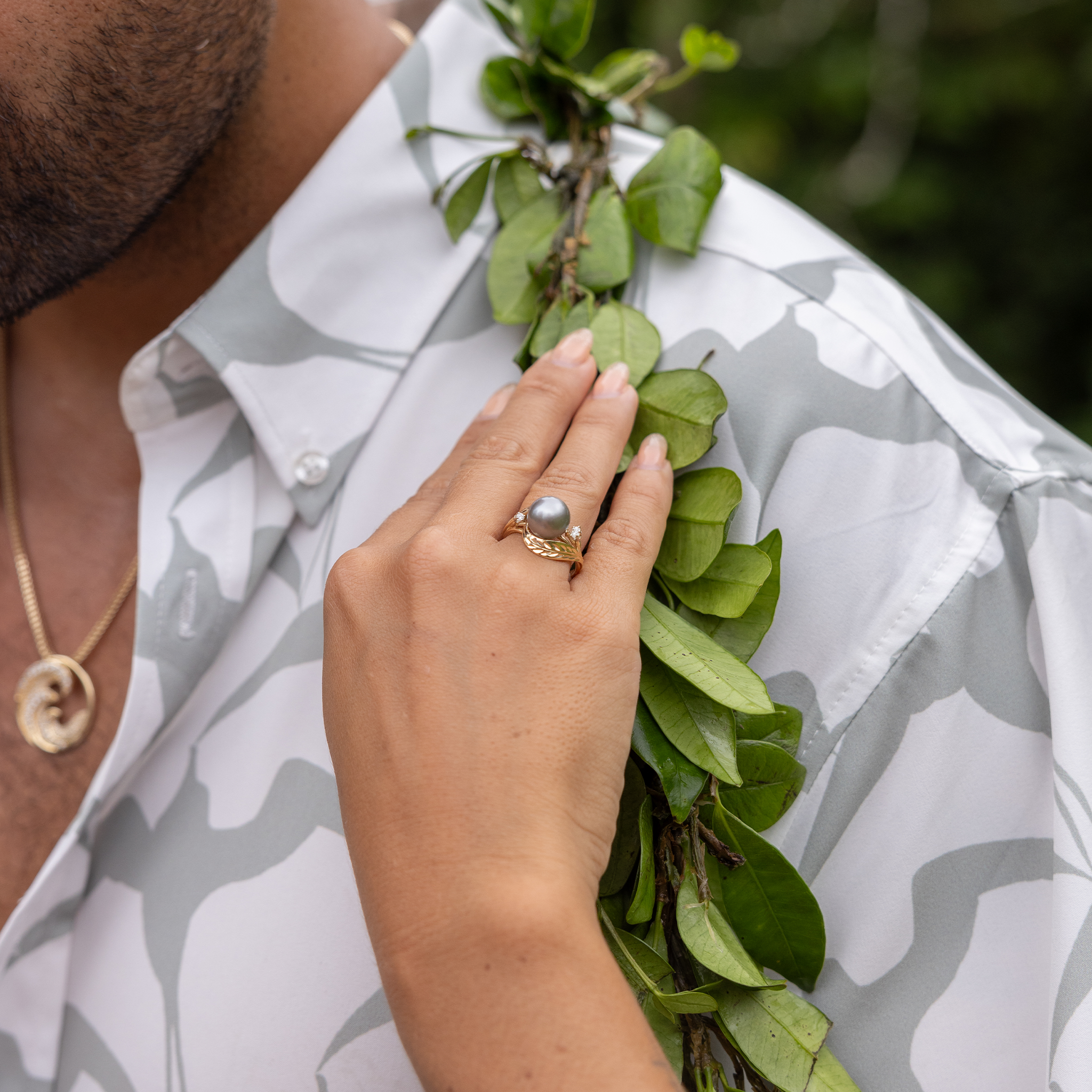 A hand with a maile leaf Tahitian black pearl ring in gold with diamonds touching a maile leaf lei resting atop a man's neck.