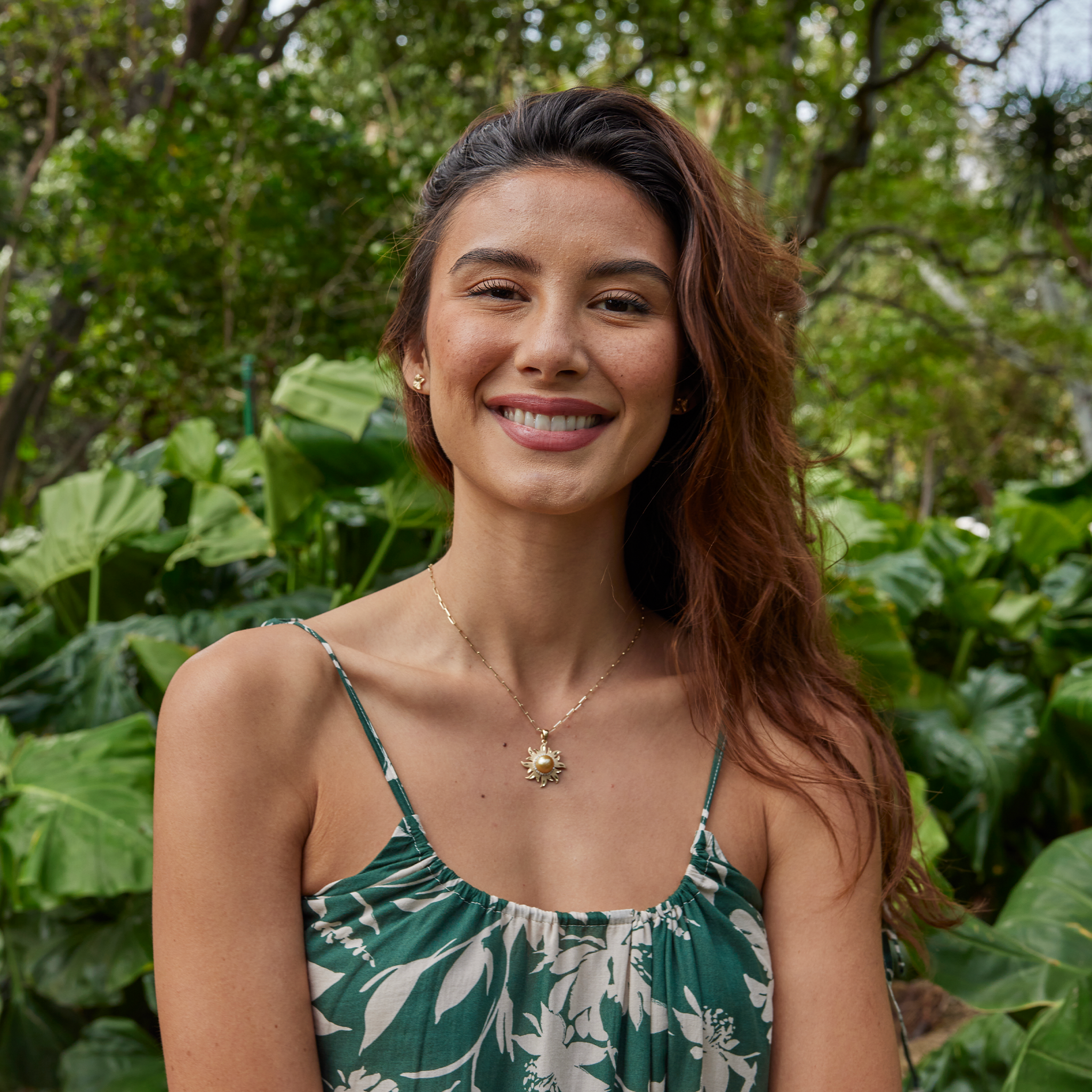 Diamond South Sea gold pearl sun pendant in 14k yellow gold on a paperclip chain, worn by a woman in a green patterned top with a plant backdrop.