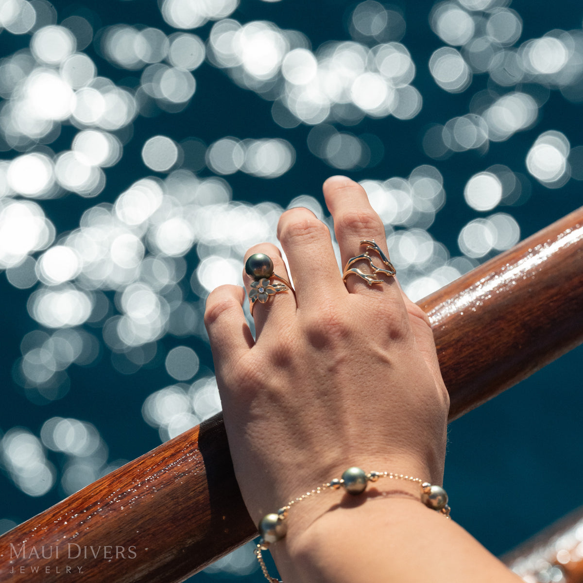 Close-up of a woman's hand wearing a gold bypass ring with a diamond plumeri and a Tahitian pearl, a gold Hertage Collection ring, and a pearl bracelet, over sparkling blue water