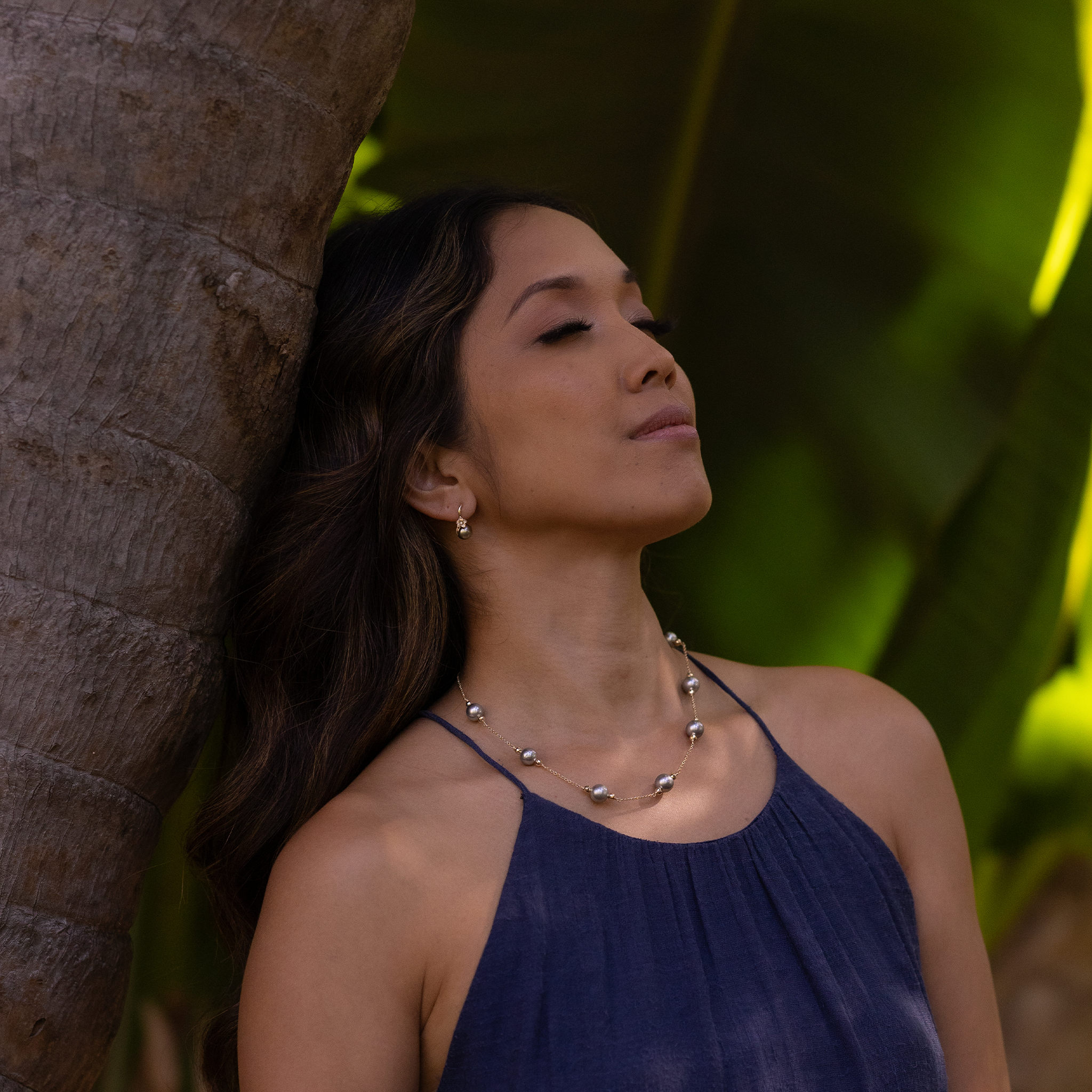A woman wearing a Tahitian black pearl necklace and plumeria earrings while leaning against a tree.