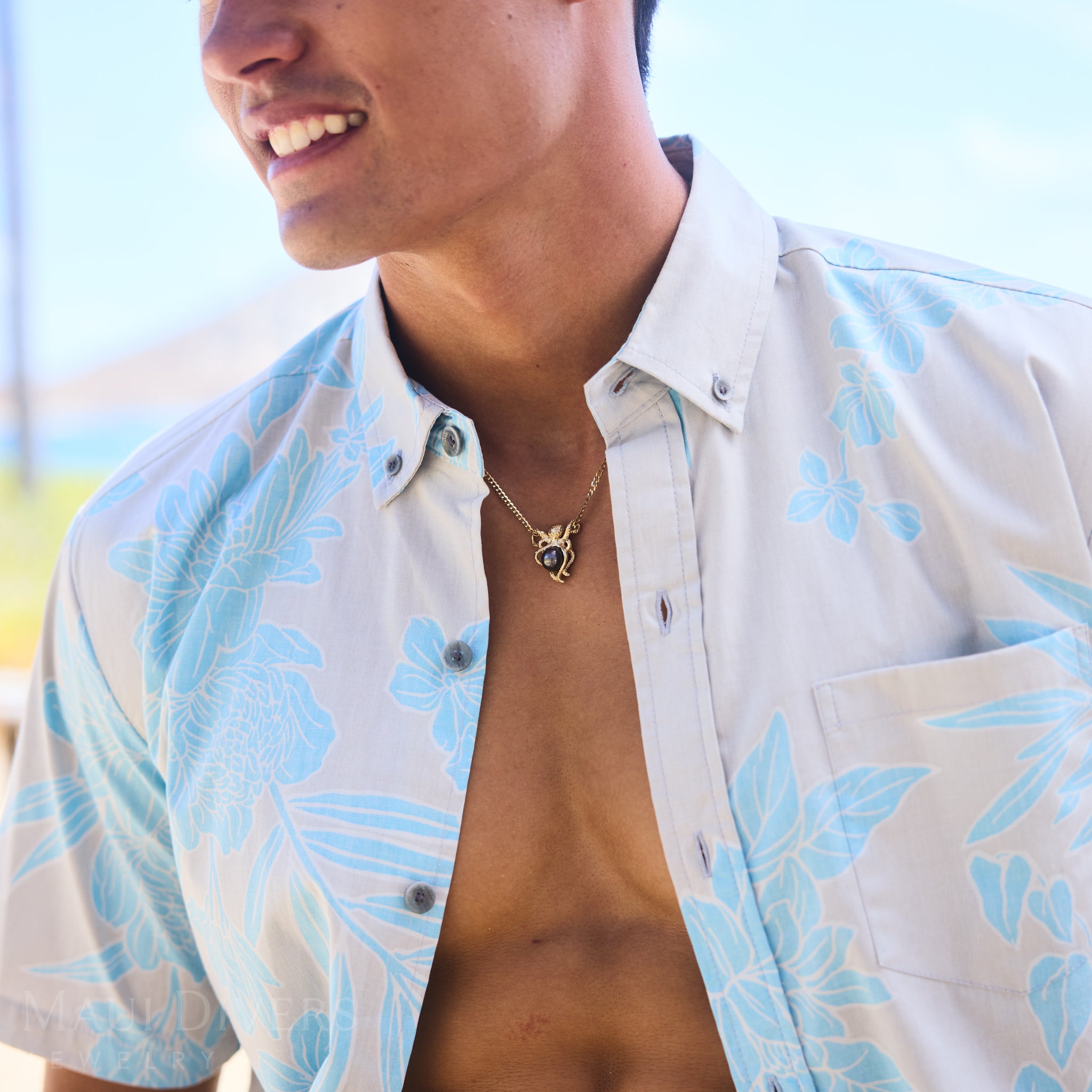 Man wearing a light blue floral shirt and a Tahitian black pearl pendant with a blurred background.