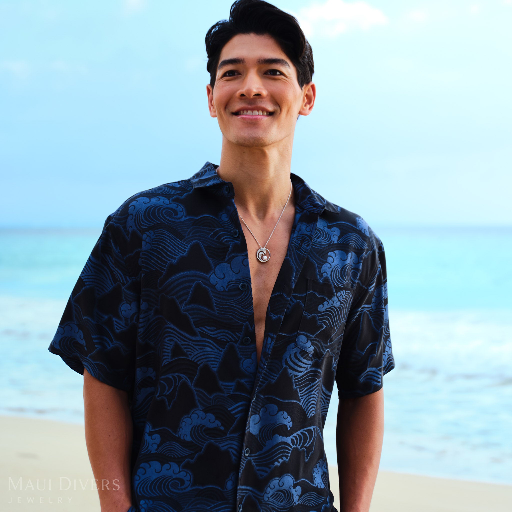 Man wearing a blue patterned shirt with a Nalu Akoya white pearl pendant in white gold, standing against a clear sky.