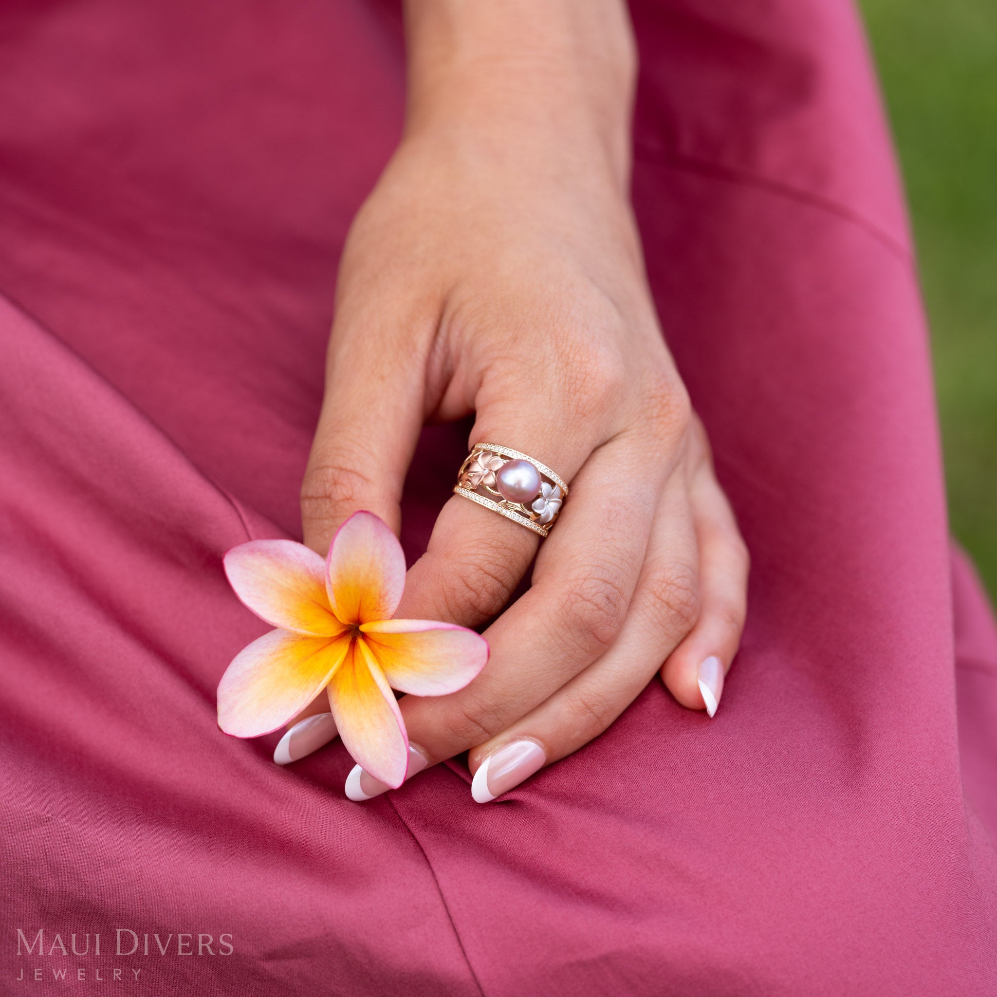 Close up of a hand wearing a Natural Lavender Freshwater Pearl Ring with Tri Color Plumerias in 14k Gold accented with Diamonds holding a light pink and yellow plumeria flower.