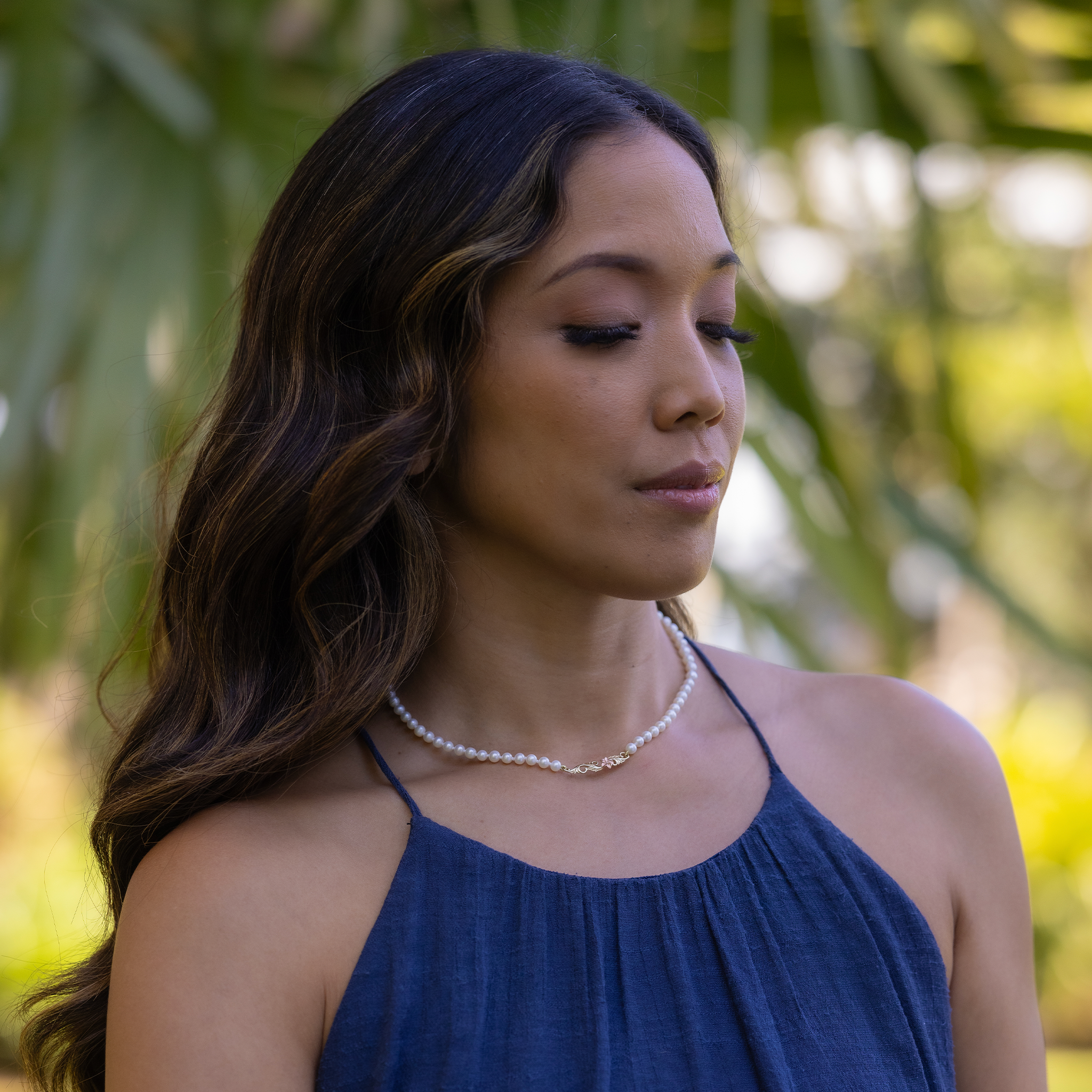 Woman in a blue top wearing the Hawaiian Heirloom Plumeria Freshwater White Pearl Necklace with blurry palm trees in the background
