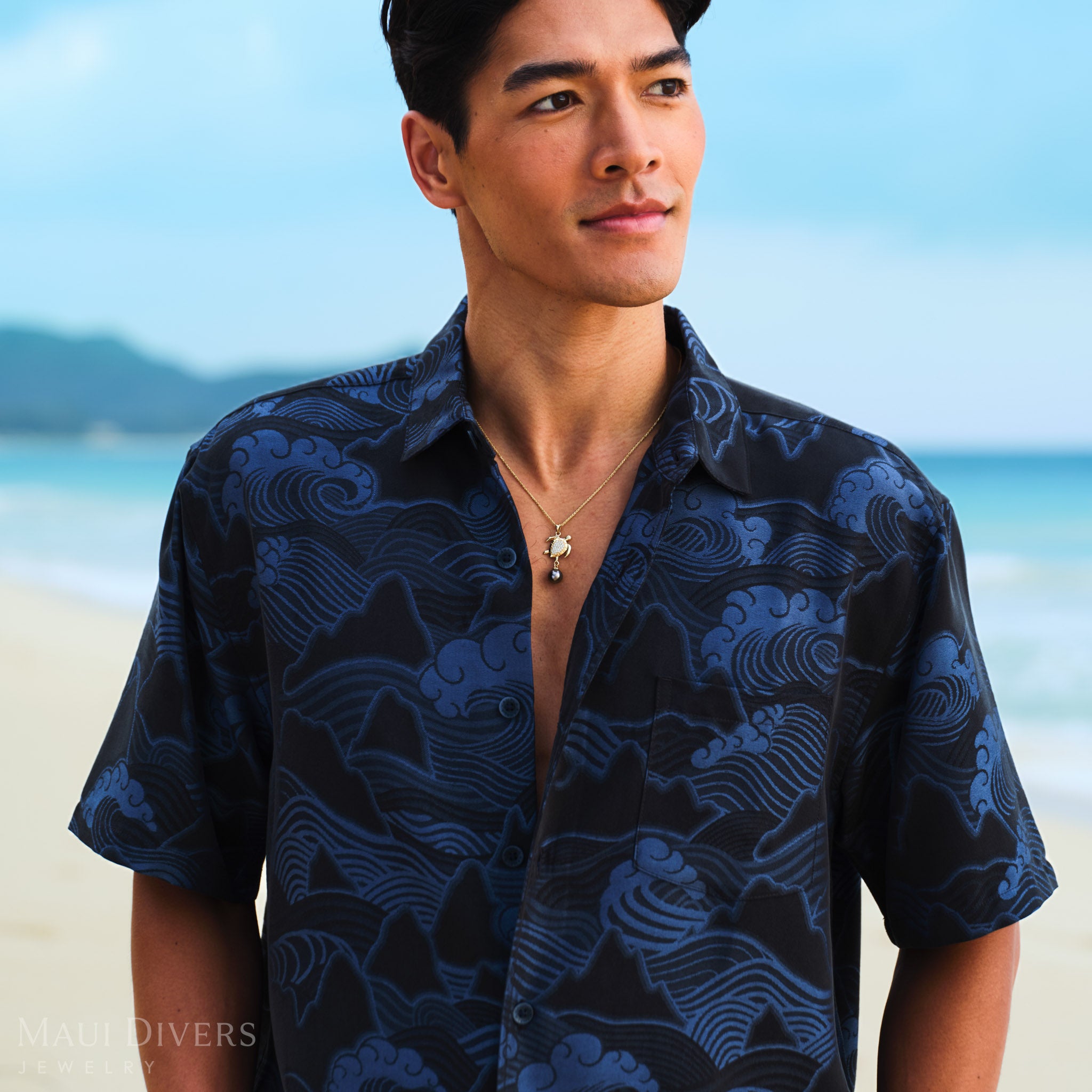 Man wearing a blue patterned shirt with a sea turtle Tahitian black pearl necklace against a blurred beach background.
