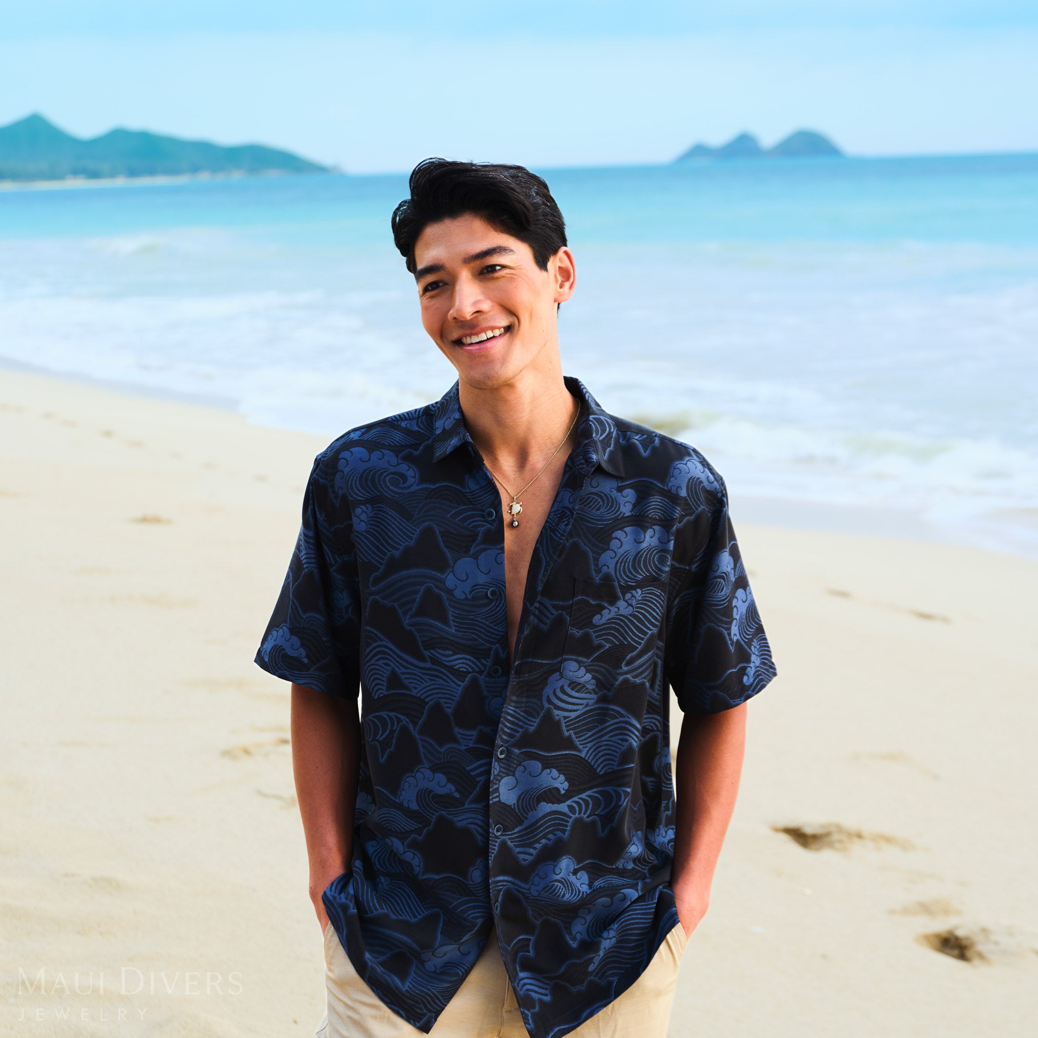 Man wearing a blue patterned shirt with a sea turtle Tahitian black pearl necklace against a blurred beach background.