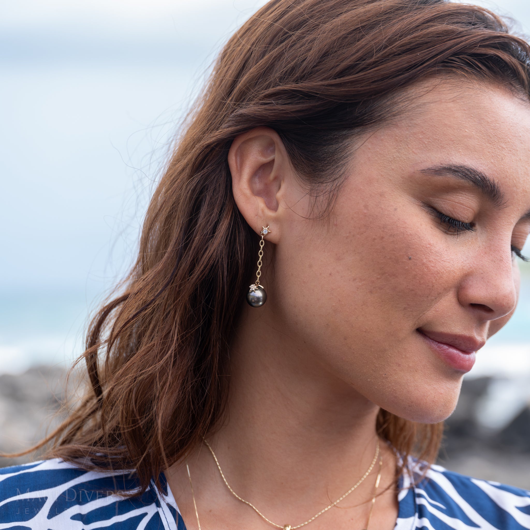 Close-up of a woman wearing the Reefs Starfish Tahitian Black Pearl Earrings in Gold with Diamonds with a blurry ocean background