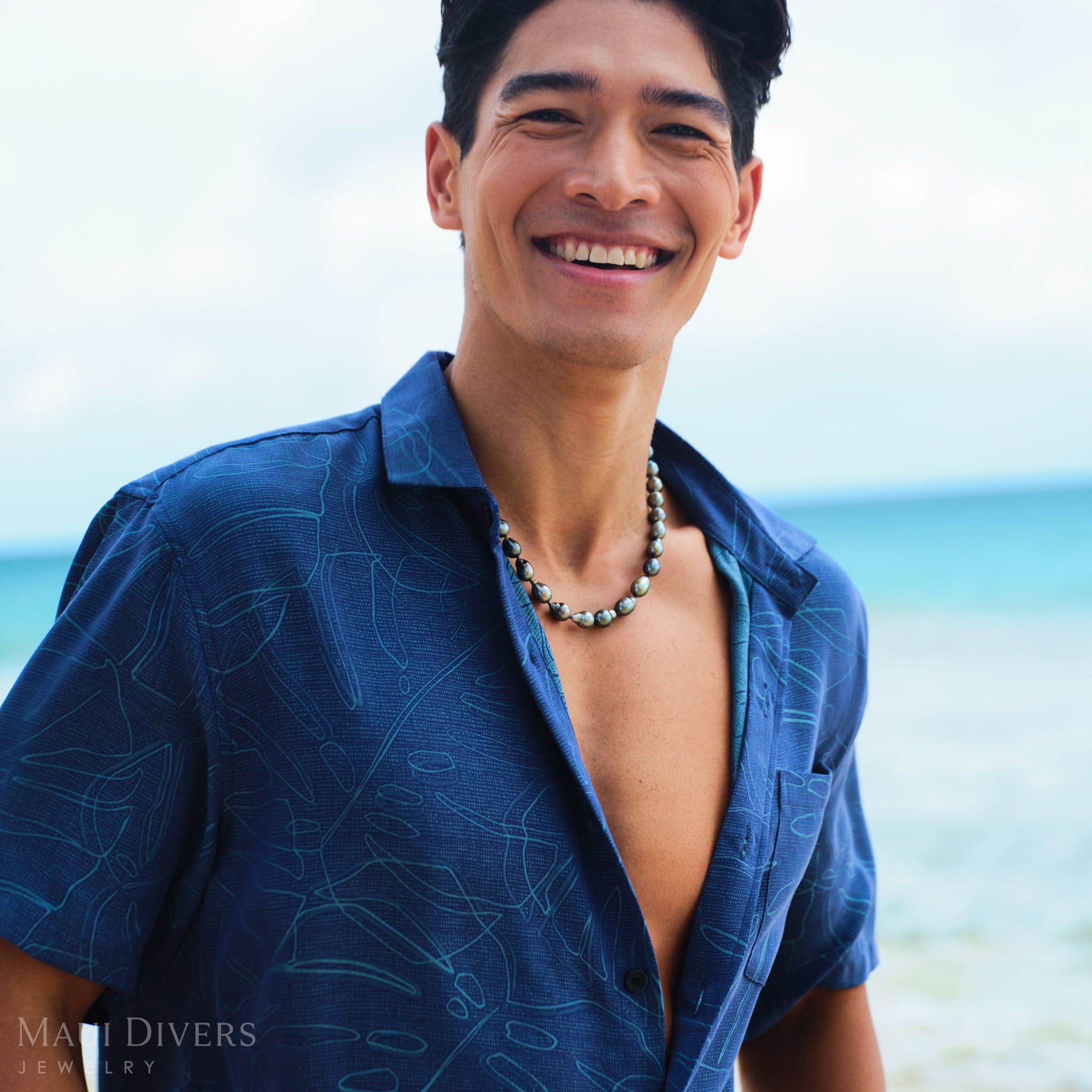 Man wearing a blue shirt with a Tahitian black pearl strand on a beach
