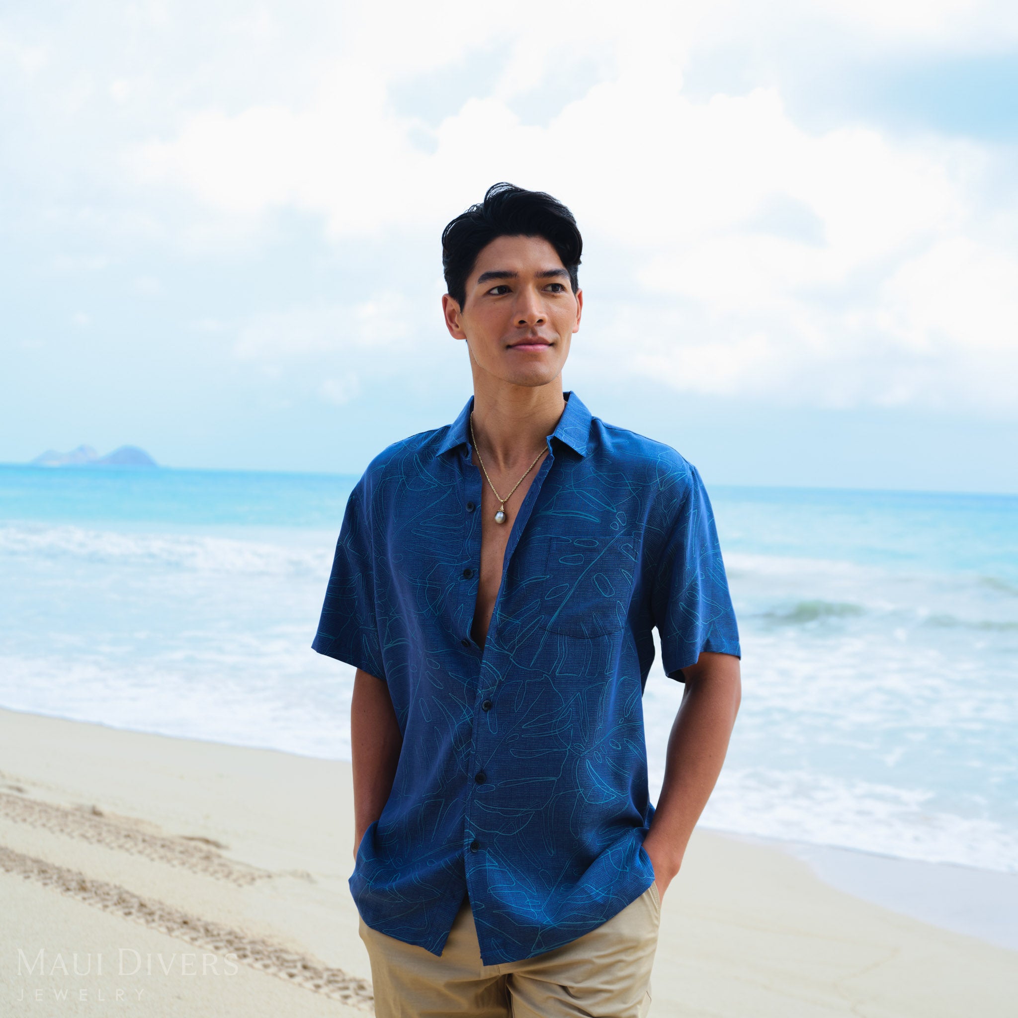 Man wearing a blue patterned shirt with a gold waterfall Tahitian black pearl pendant, set against a blurred beach background.