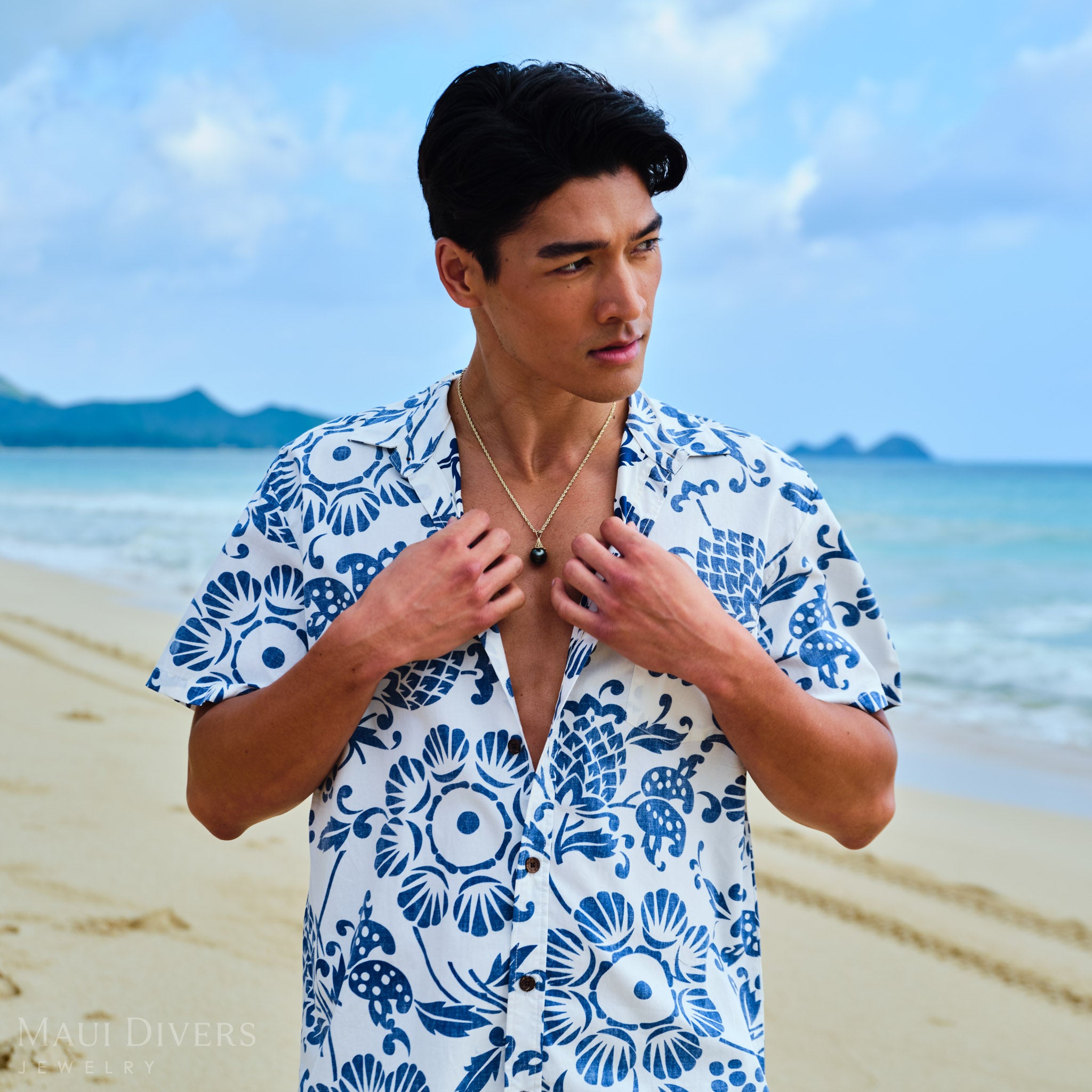 Man wearing a blue and white patterned shirt with a gold fish net Tahitian black pearl necklace on a beach with ocean and sky in the background