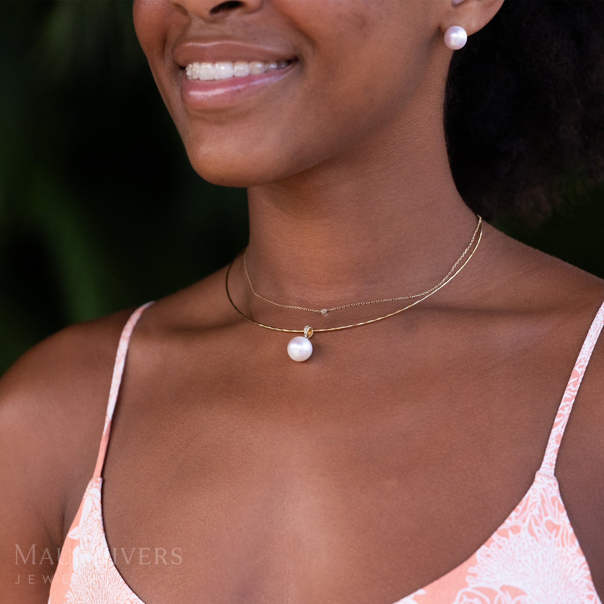 A close-up of a smiling woman in a pastel colored top wearing a Freshwater White Pearl Pendant in 14k yellow gold with diamonds on an omega chain, layered with a tiny diamond heart necklace, against a blurred leafy green background