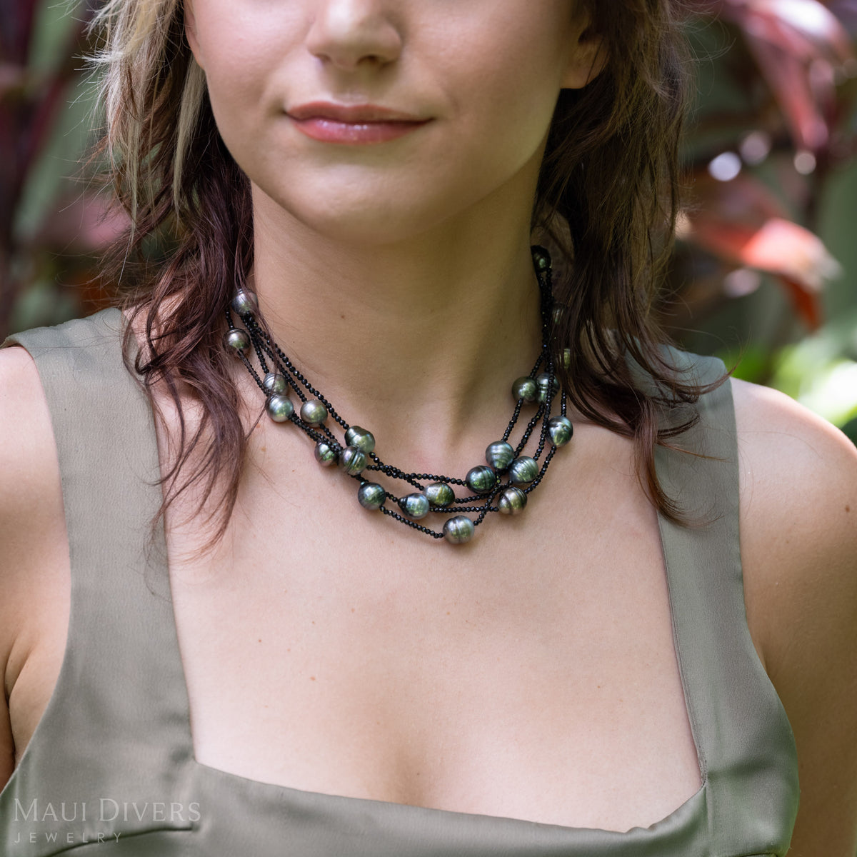 A close-up of a woman wearing a multi-layer Tahitian black pearl and black spinel necklace. She is wearing an olive green satin top with a blurred tropical garden background.