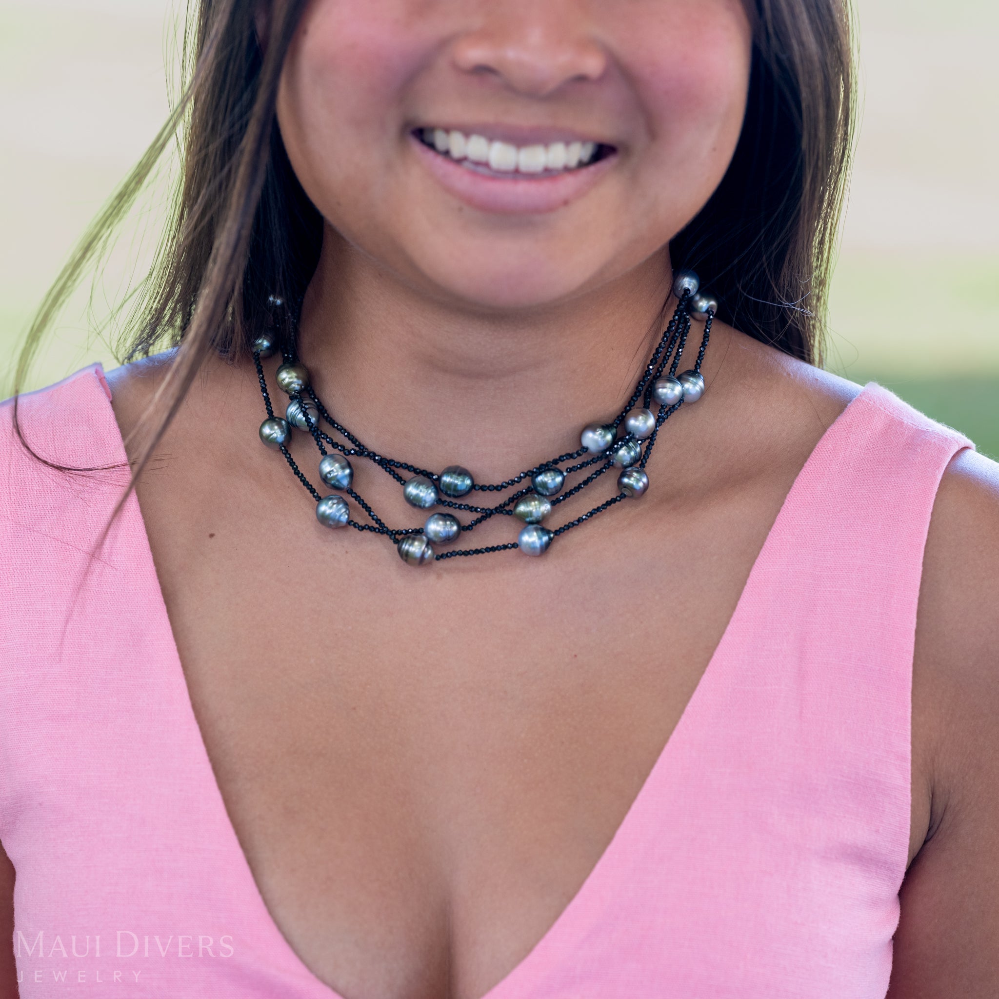 A close-up of a smiling woman in a light pink top wearing a Tahitian Black Pearl and Black Spinel Layered Necklace with a 14k yellow gold clasp, against a blurred green grass background