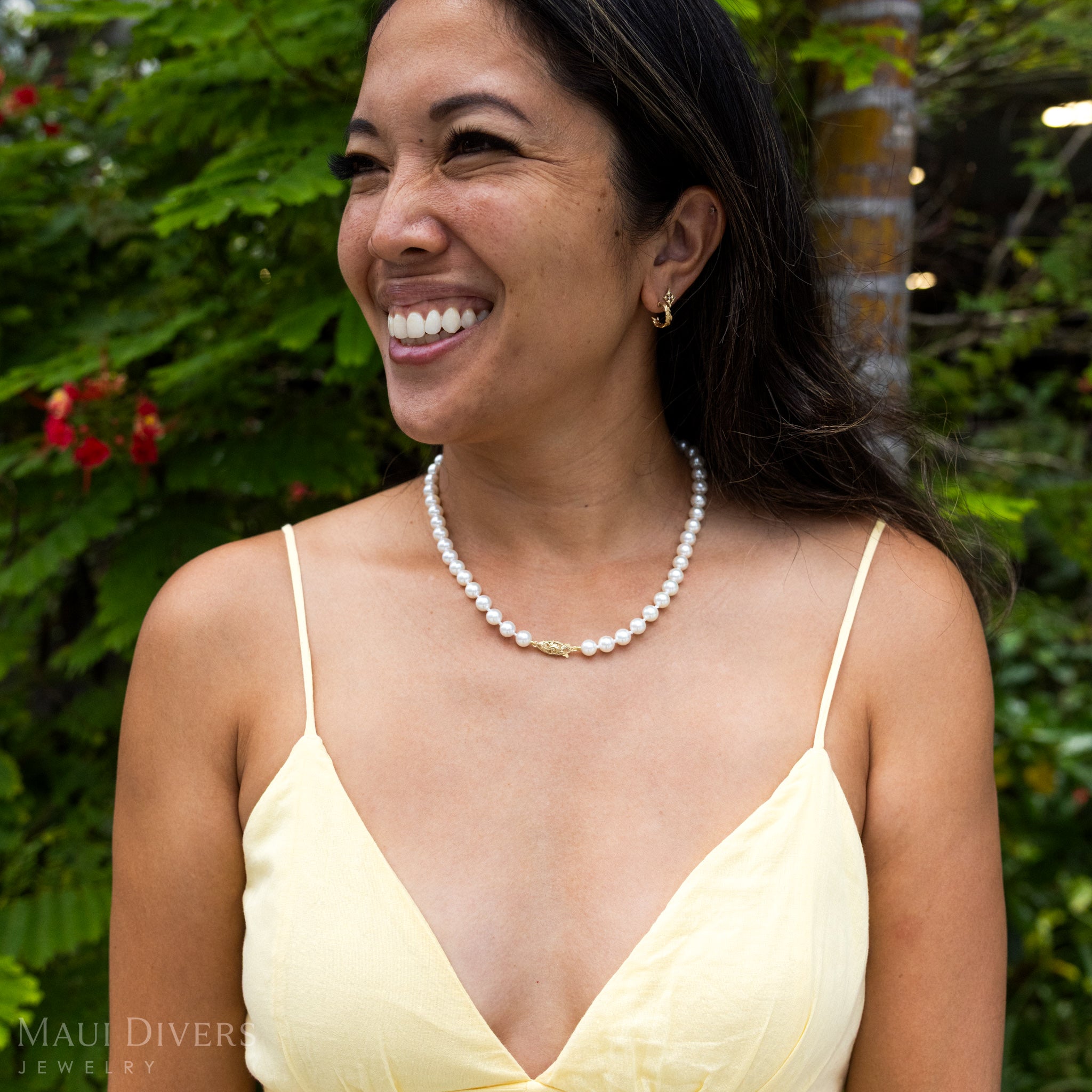 Smiling woman in a yellow top wearing an Akoya White Pearl Strand with 14k yellow gold Living Heirloom clasp against a blurred leafy green background