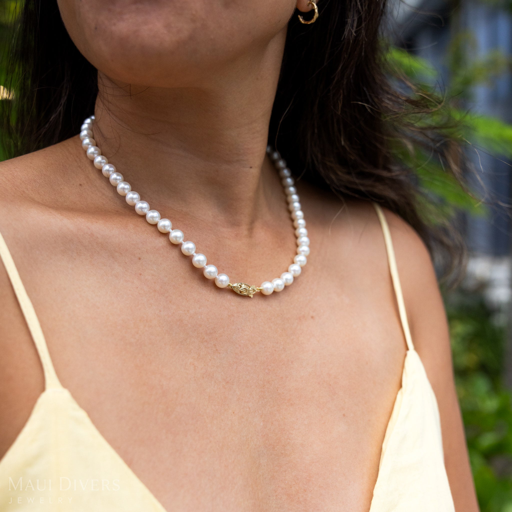 Close-up of a woman in a yellow top wearing an Akoya White Pearl Strand with 14k yellow gold Living Heirloom clasp against a blurred leafy green background