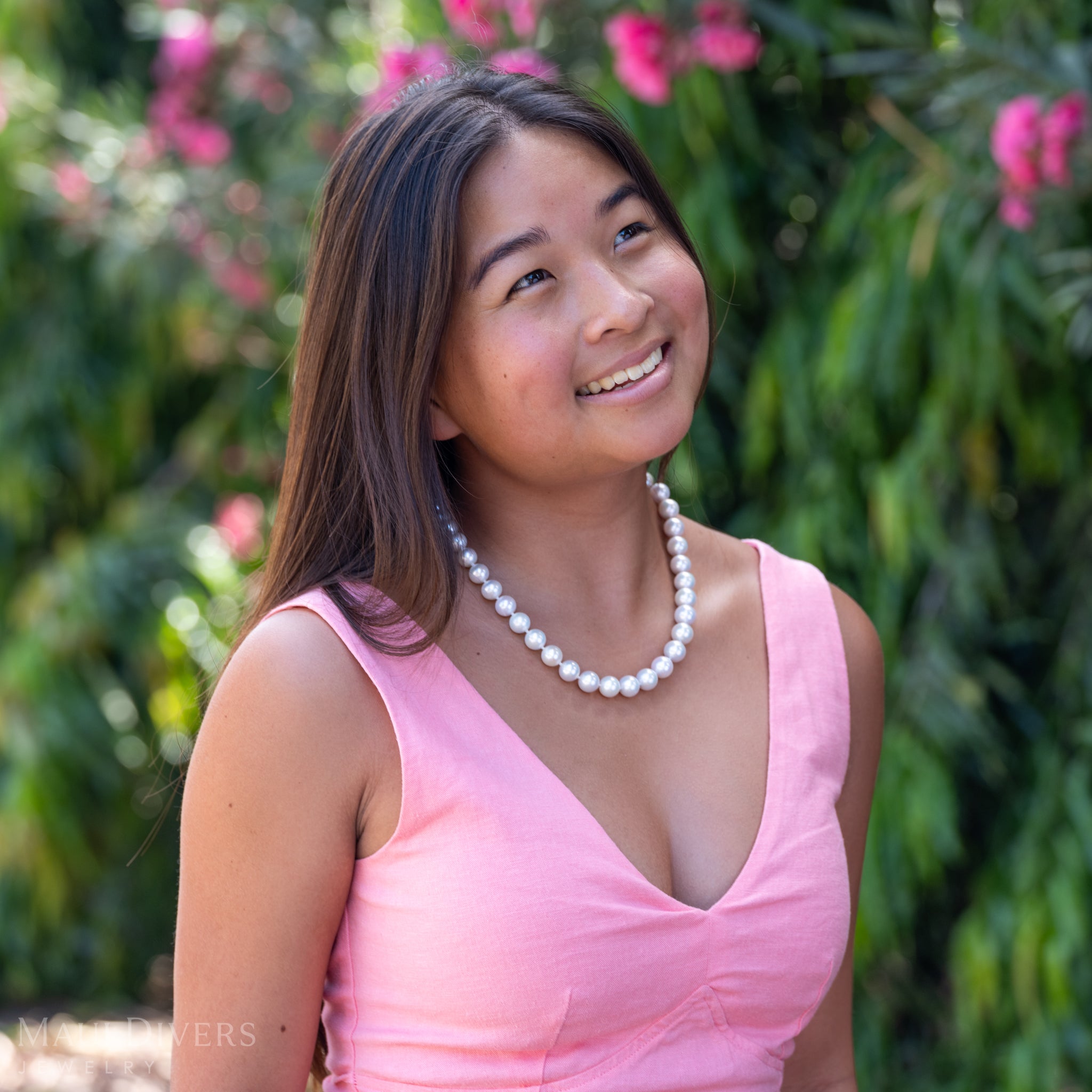 A smiling woman in a light pink top wearing a Freshwater White Pearl Strand with Diamond Plumeria Clasp in 14k Yellow Gold, against a blurred leafy green and pink floral background