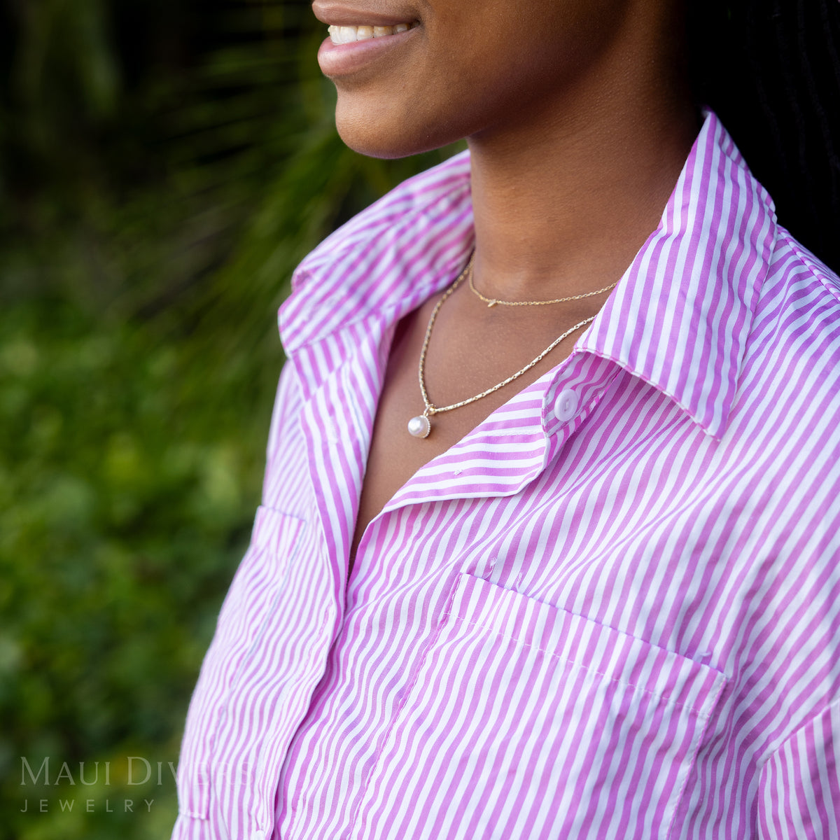 A side view close-up of a smiling woman in a pink and white striped top wearing a Halo Akoya White Pearl Pendant in 14k yellow gold with diamonds on a raso chain, layered with a tiny diamond heart necklace, against a blurred leafy green background