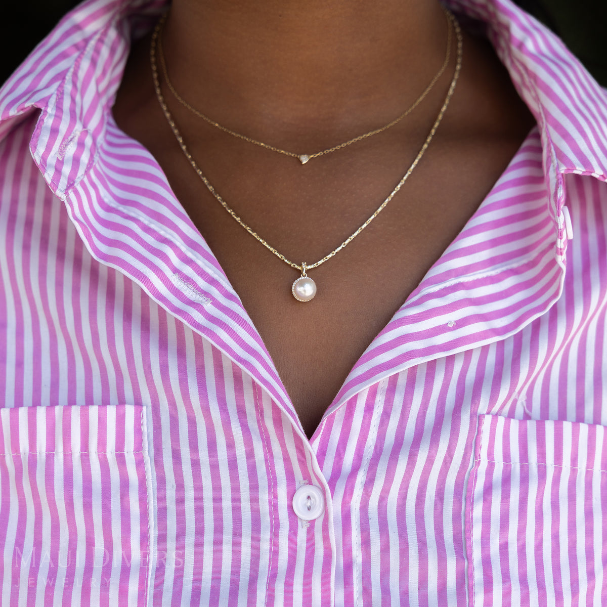 A close-up of a woman in a pink and white striped top wearing a Halo Akoya White Pearl Pendant in 14k yellow gold with diamonds on a raso chain, layered with a tiny diamond heart necklace