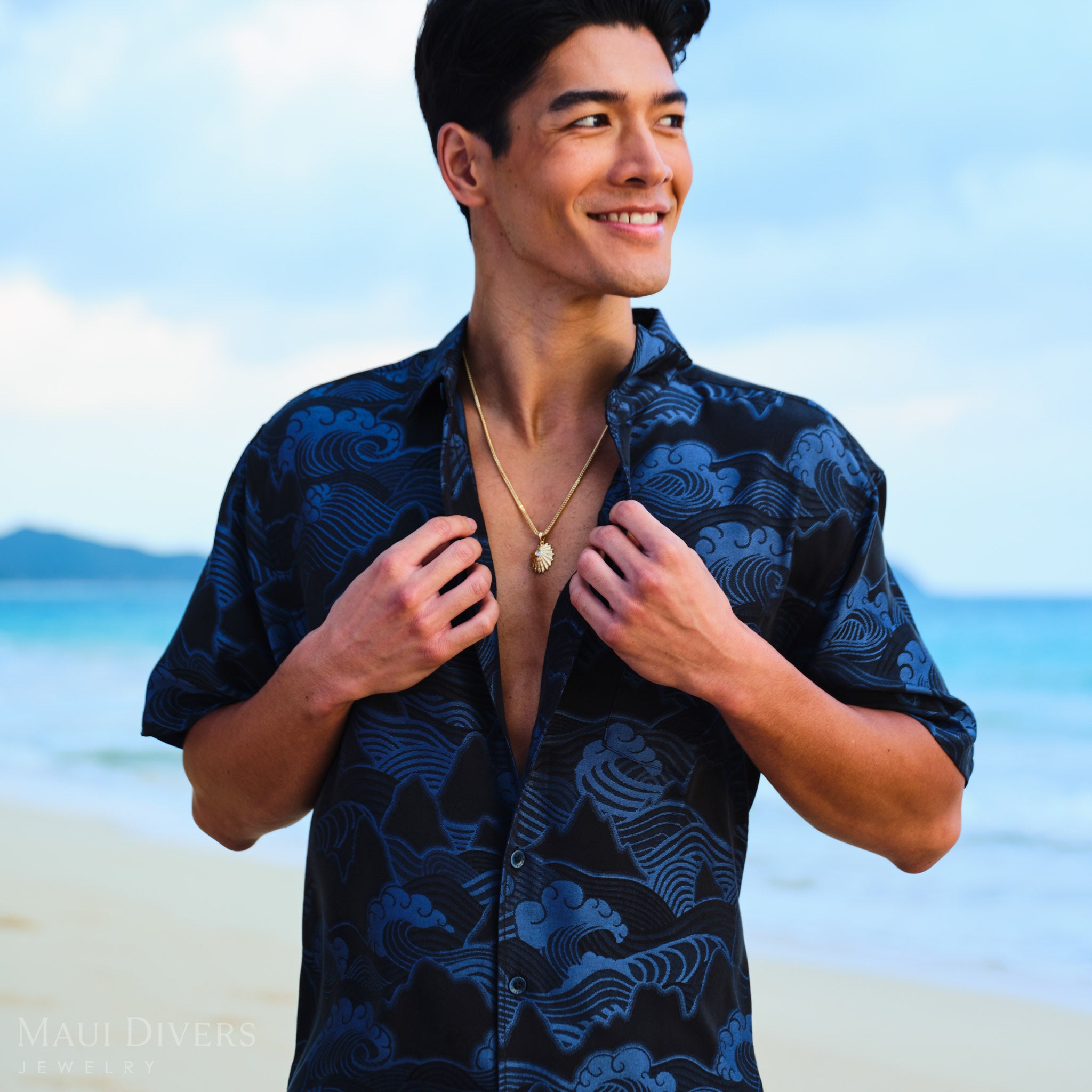 Man wearing a dark blue patterned shirt and an ʻopihi pendnat on a beach with ocean and sky in the background