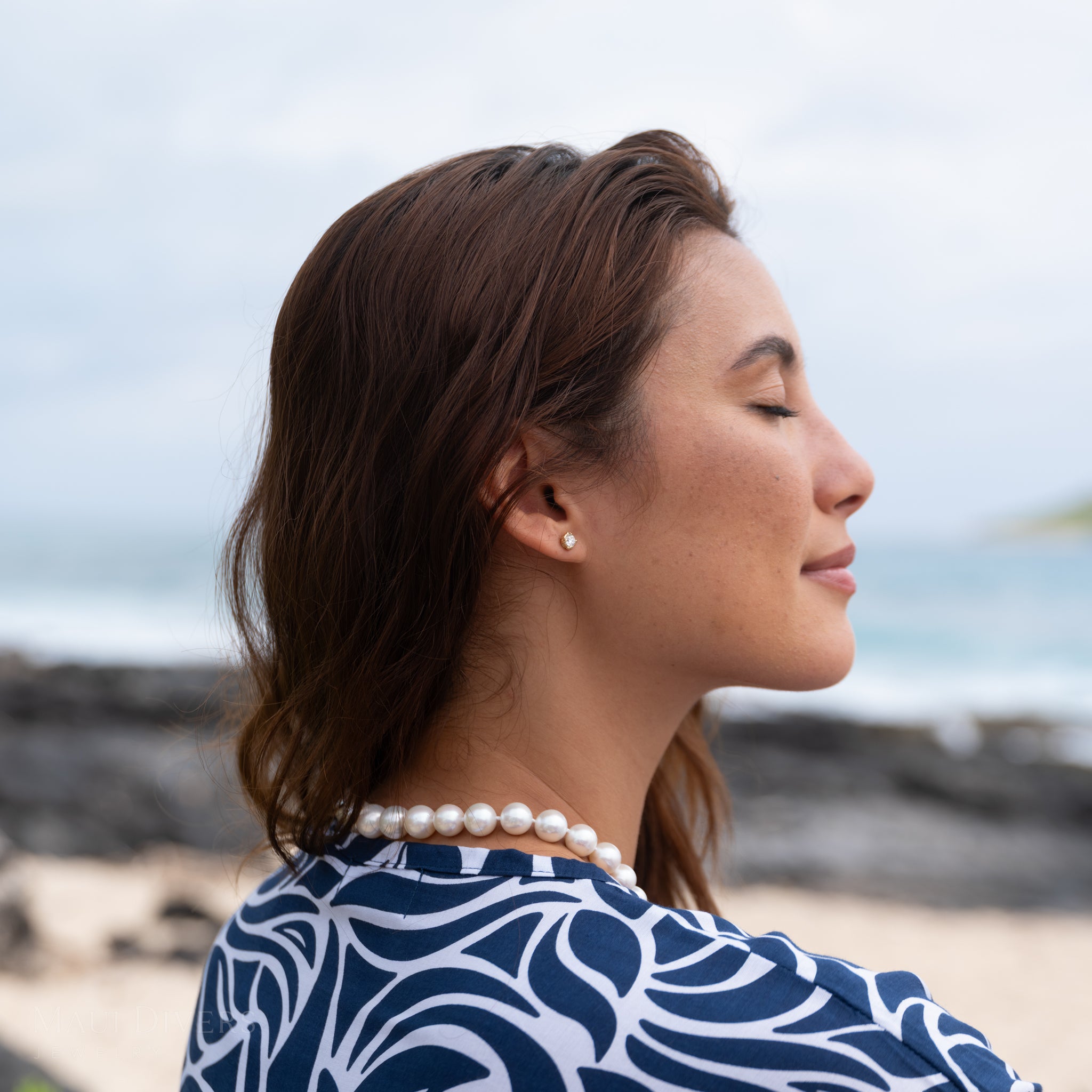 Woman wearing a Hidden Hawaiʻi Maile Lab Grown Diamond Earring and white pearl necklace with a blurred beach background