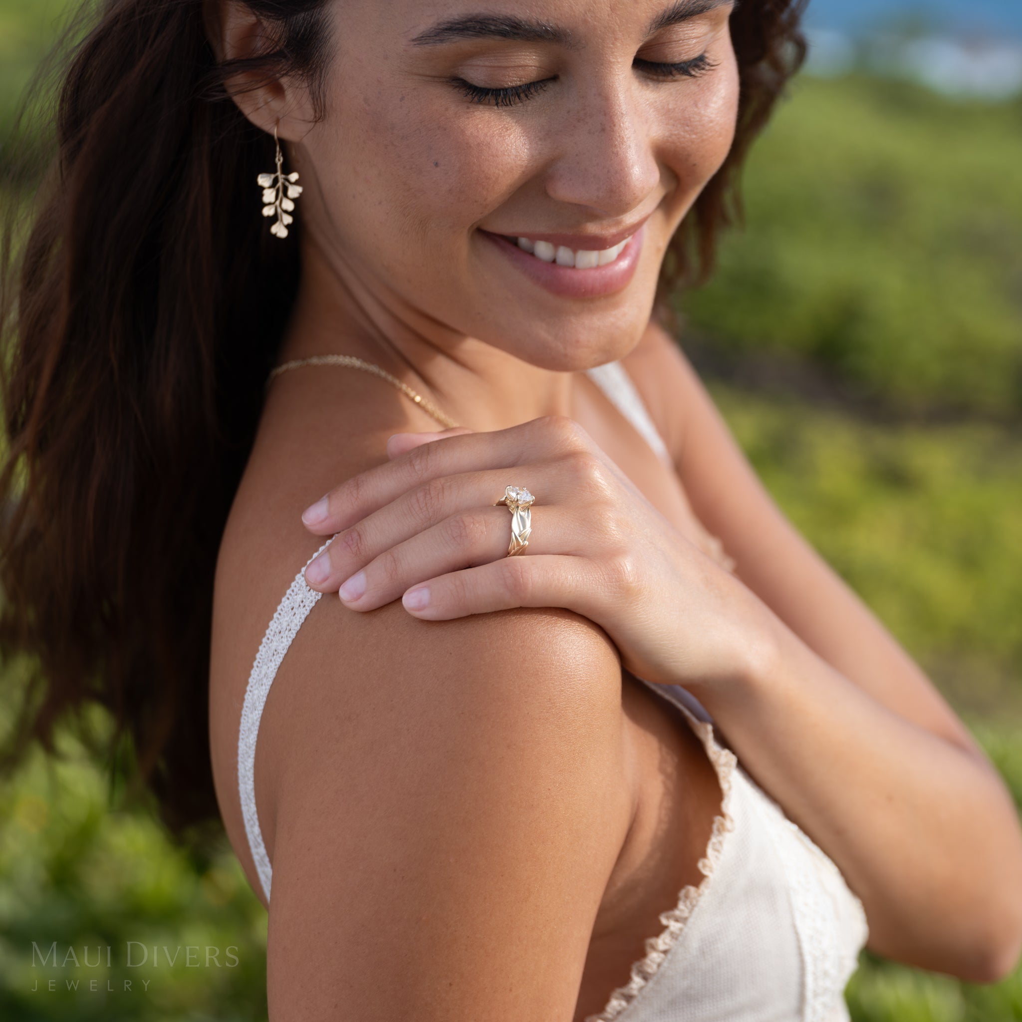 Woman wearing a gold maile lab-grown diamond engagement ring outdoors with greenery in the background