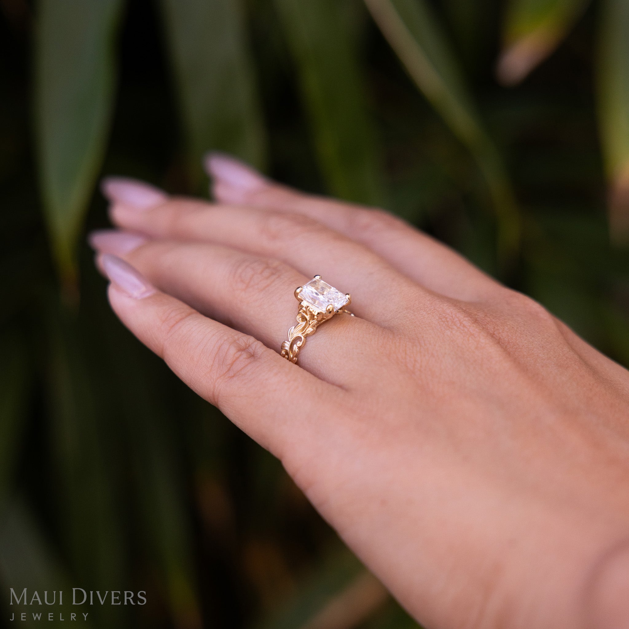 Close-up of a Living Heirloom Solitaire Lab Grown Diamond Engagement Ring in 14k yellow gold worn on the ring finger of a hand, against a blurred leafy green background