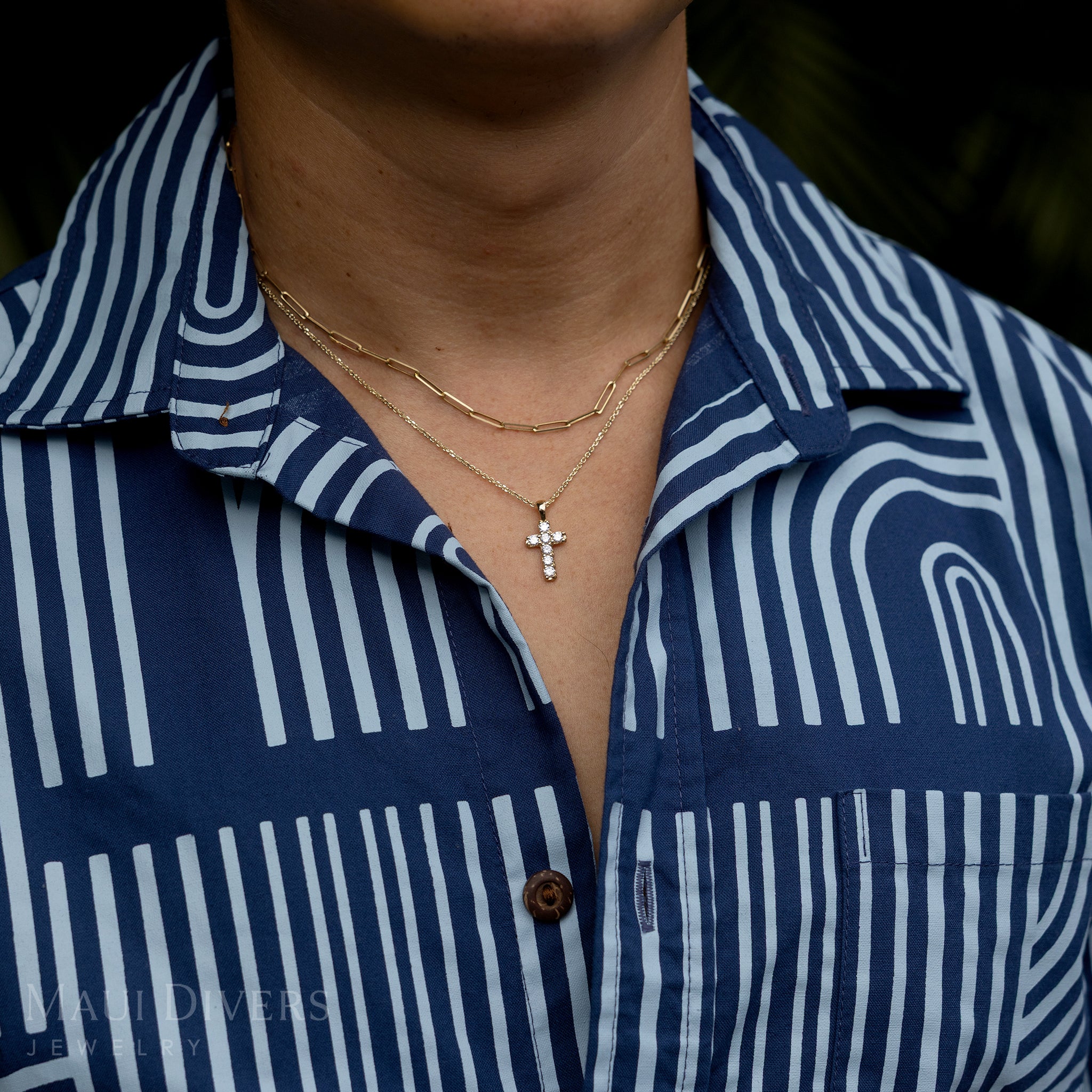 A close-up of a guy in a two-toned blue patterned shirt wearing a Hidden Hawaiʻi Maile Lab Grown Diamond Cross Pendant in 14k yellow gold around his neck, layered with a paperclip chain against a blurred green leaf background