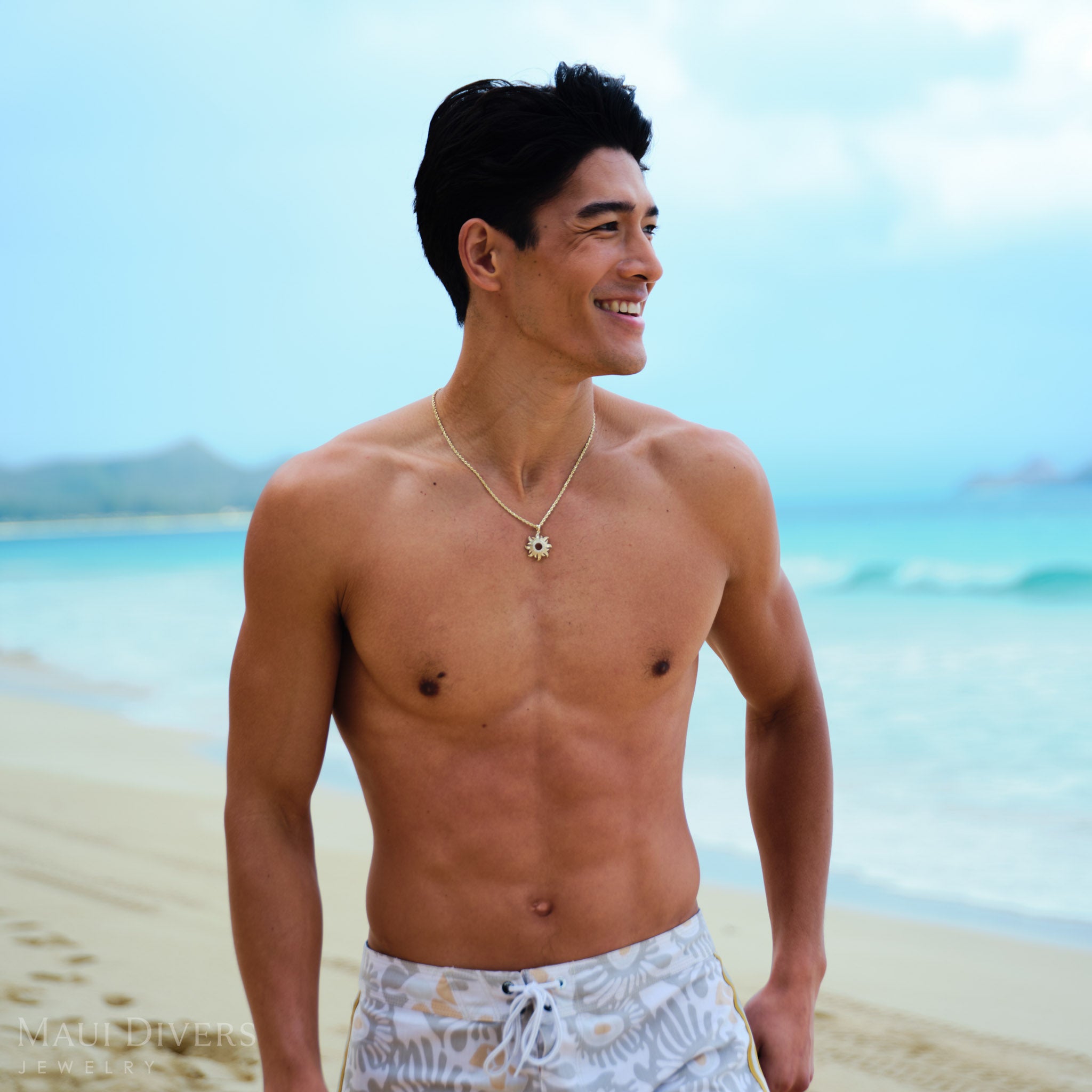 Man standing on a beach wearing a gold sun pendant on a rope chain with clear blue water and sky in the background