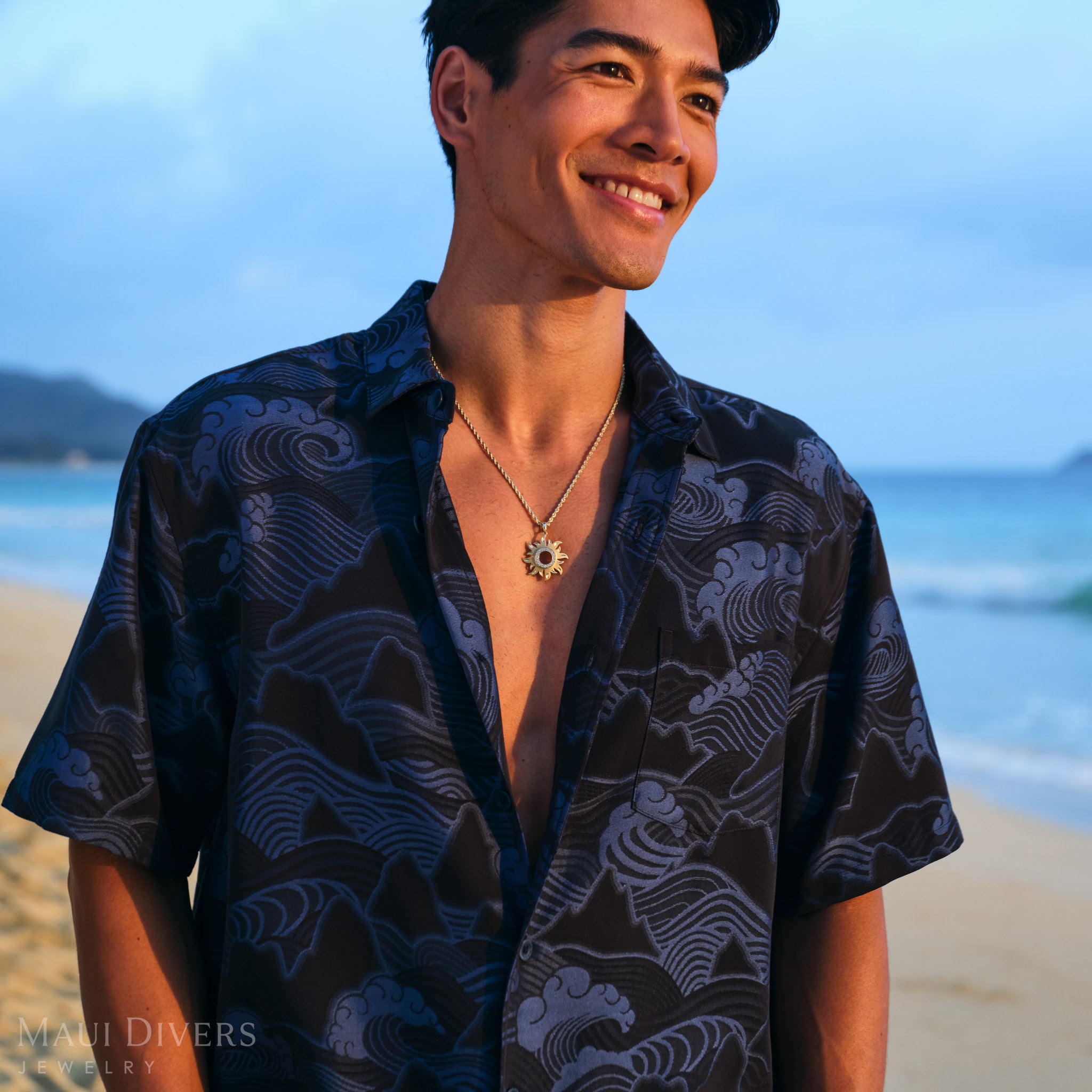Man on a beach wearing a dark blue patterned shirt with a gold sun pendant on a rope chain