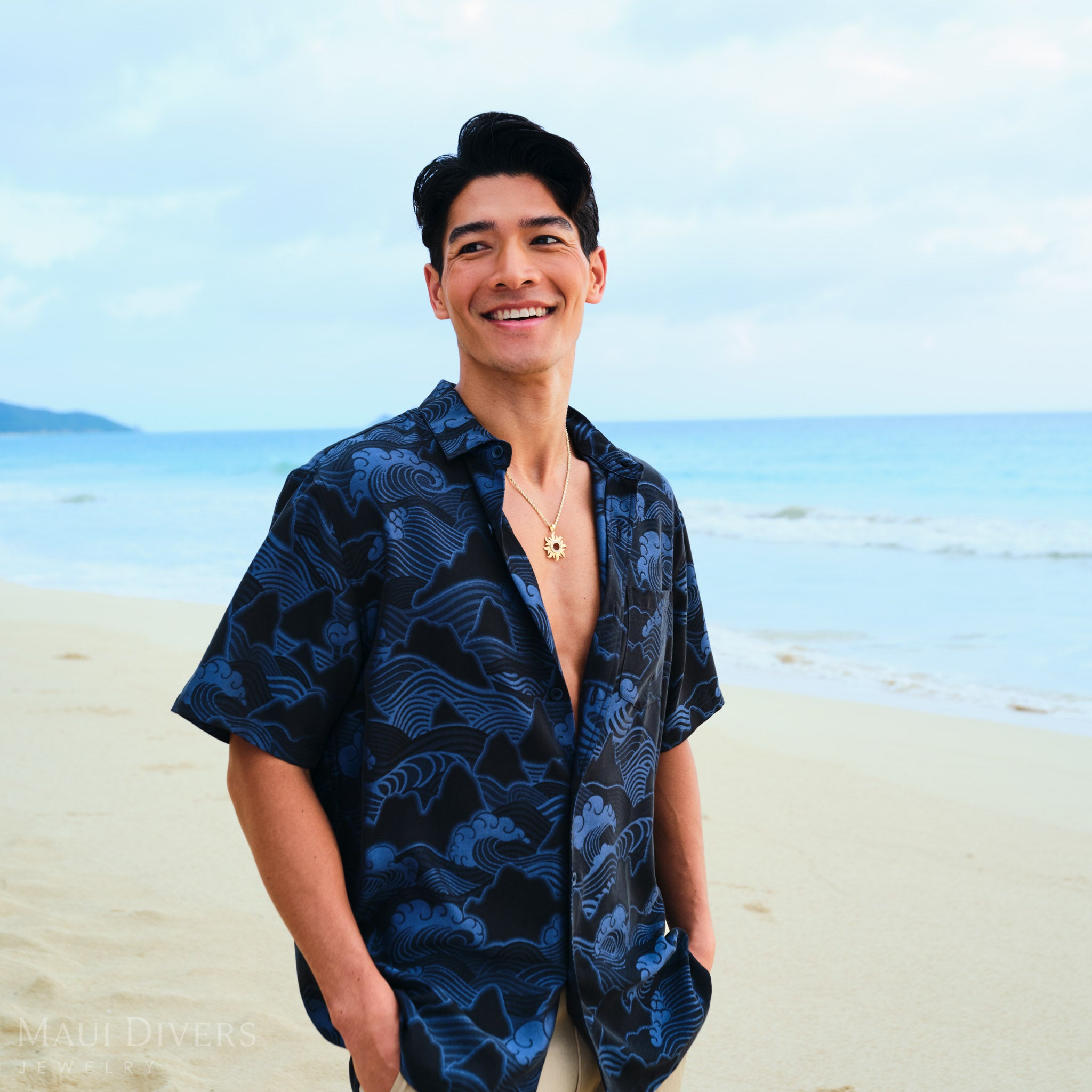 Man on a beach wearing a dark blue patterned shirt with a gold sun pendant on a rope chain