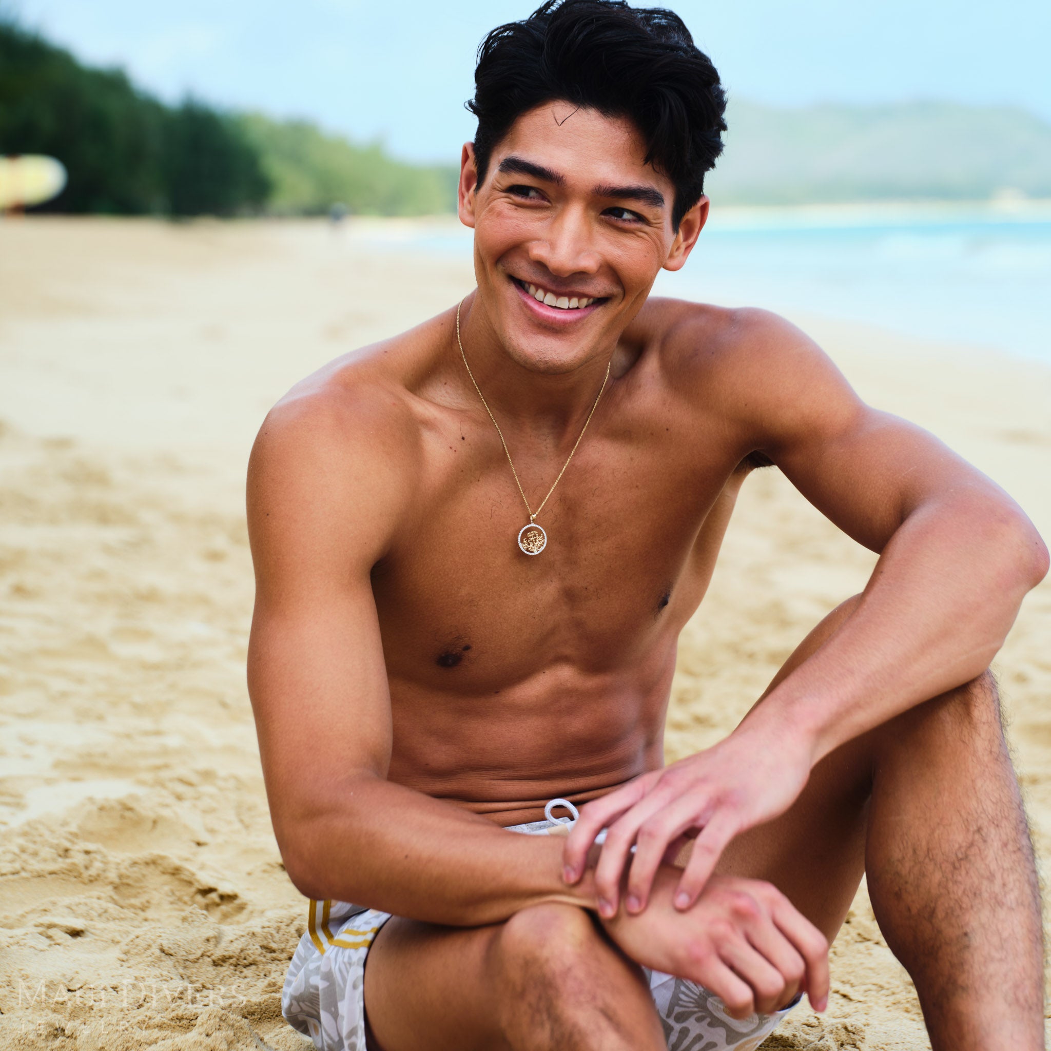 Man sitting on a beach wearing a gold coral branch pendant on a rope chain with a clear sky and water in the background