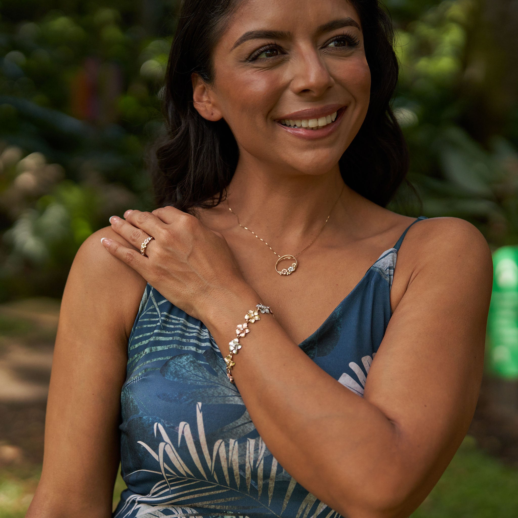 Woman in a lush garden wearing matching plumeria necklace, ring and bracelet in tri-color gold