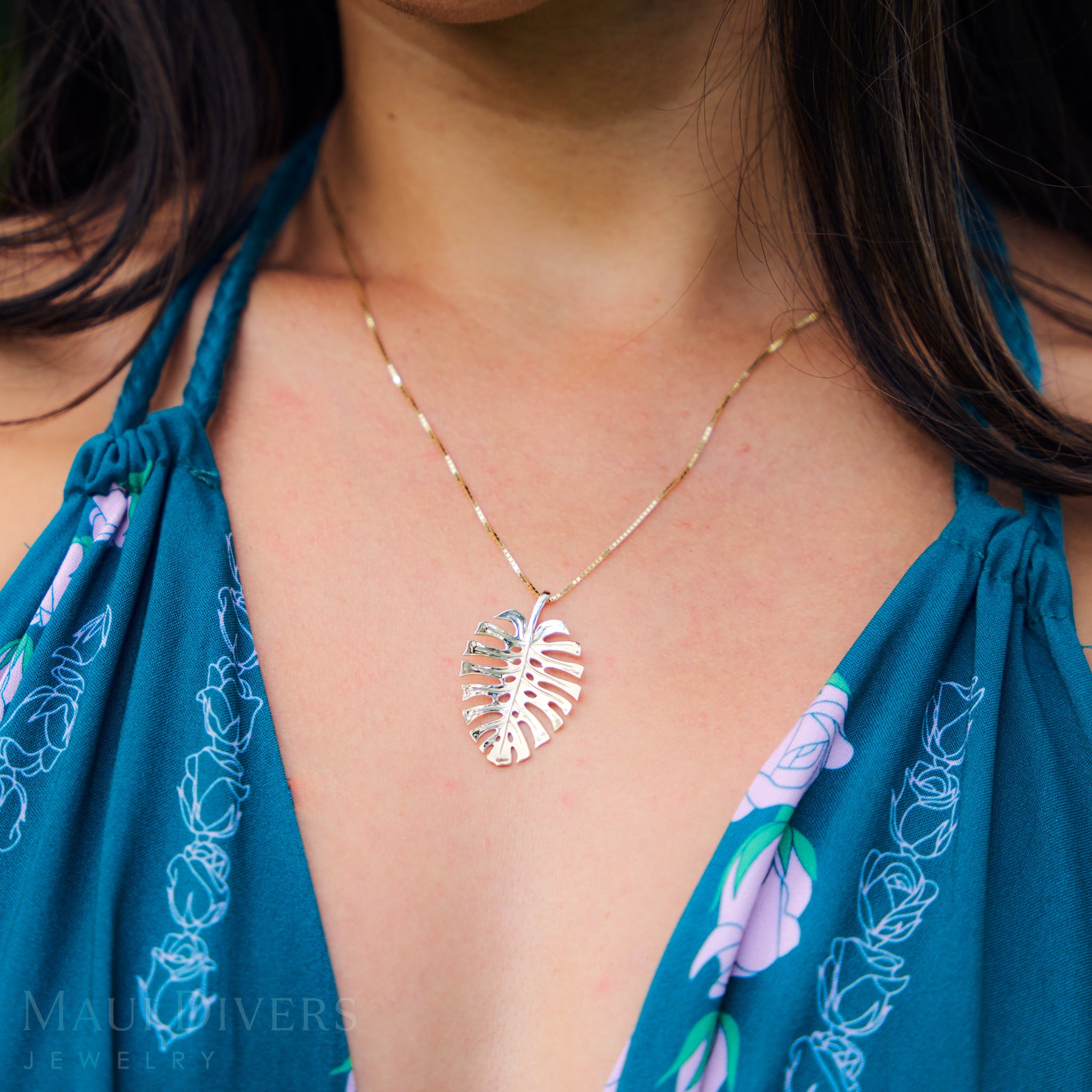 Gold Monstera leaf necklace worn by a person in a blue floral dress