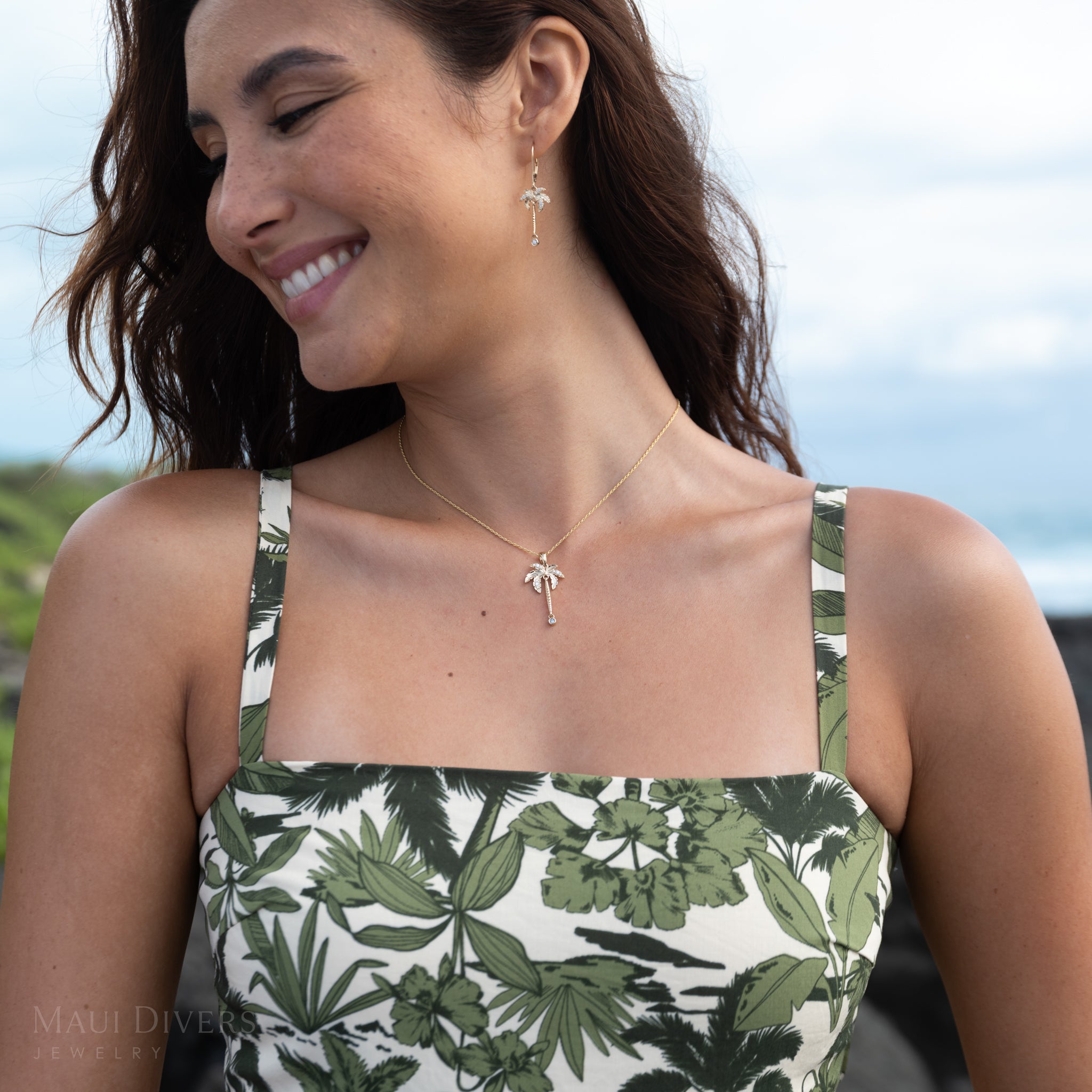 Close-up of a woman wearing a gold and diamond palm tree pendant on a rope chain, matching earrings, and a patterned white and green dress with a sky in the background