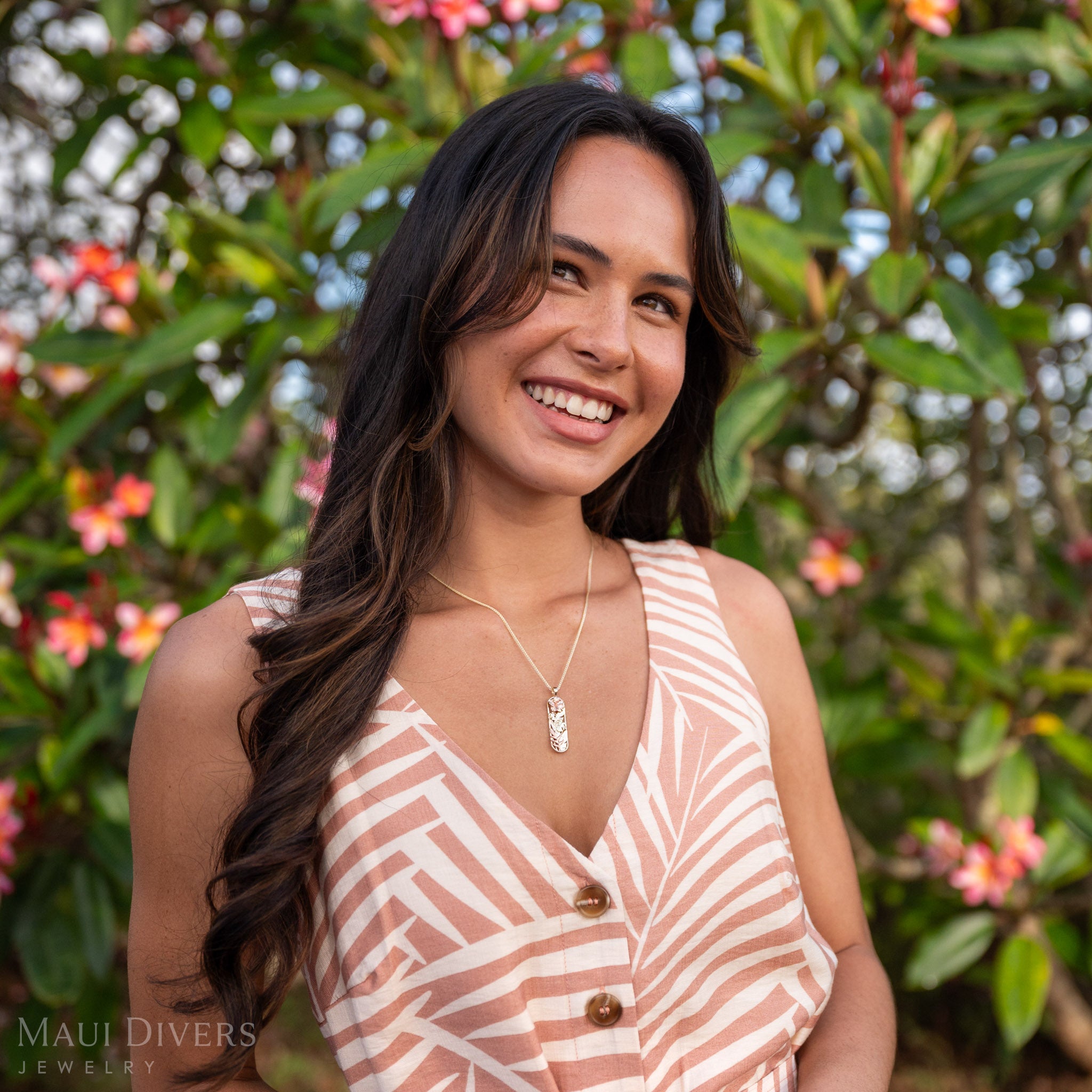 Woman smiling and wearing a 14k Tri-Gold Pendant featuring heliconia, monstera, and hibiscus flowers against a blurred background of green leaves and pink and white flowers.