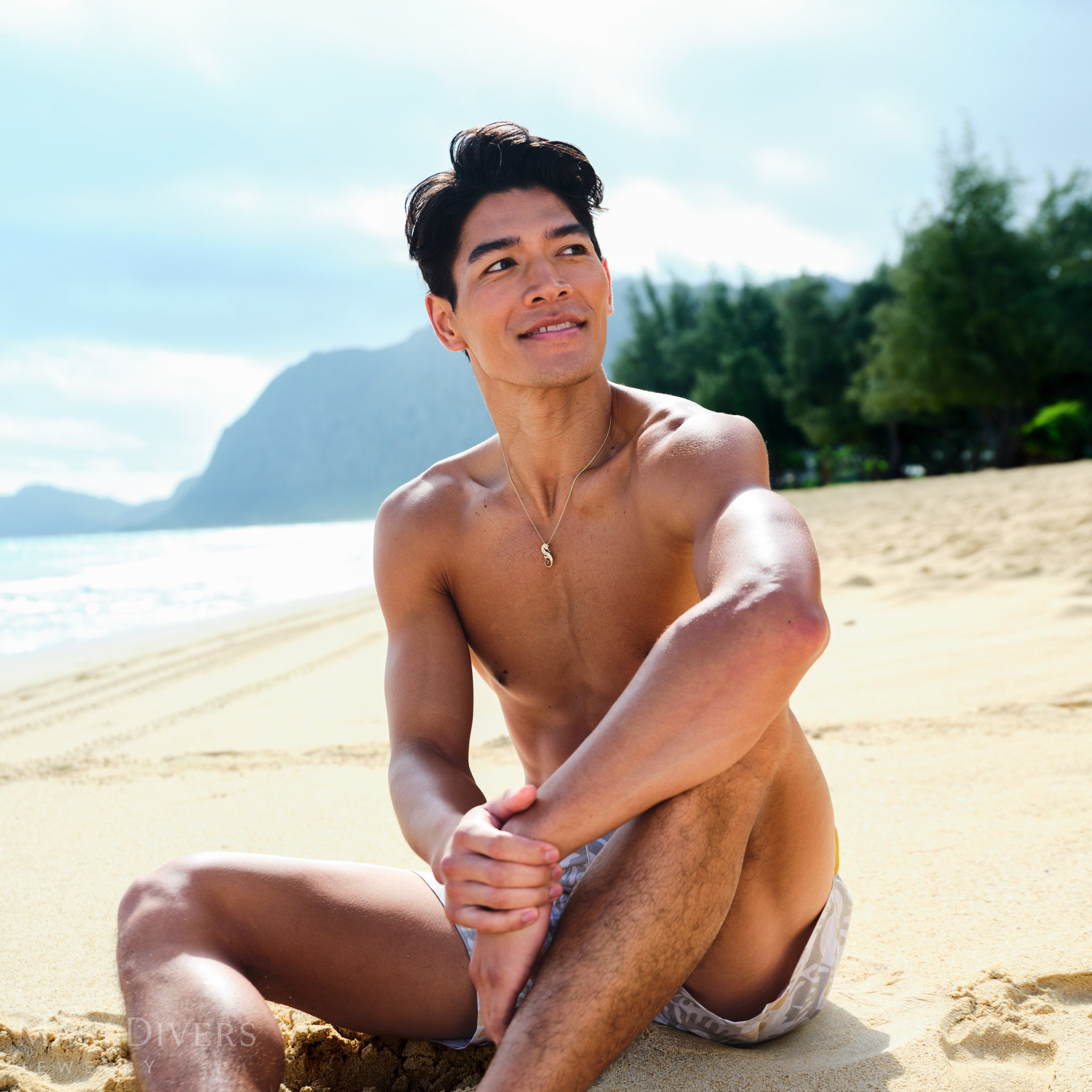 Man sitting on a beach wearing a gold seahorse necklace with mountains and trees in the background