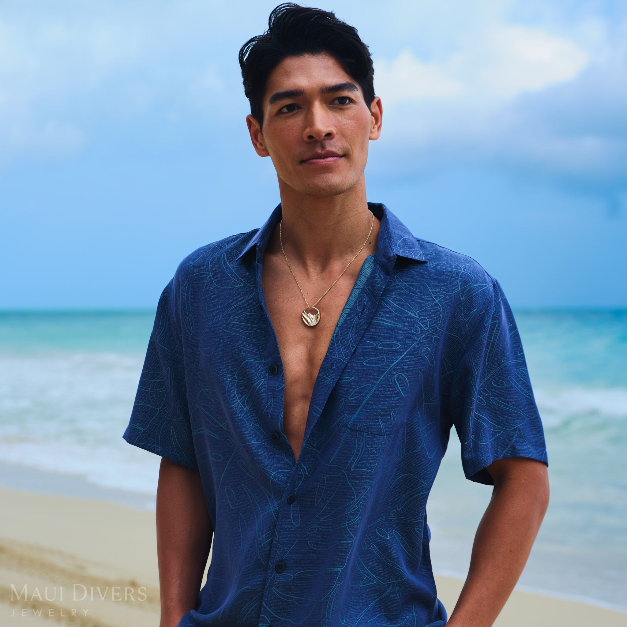 Man wearing a gold Koʻolau mountain pendant and a patterned monstera shirt on a beach