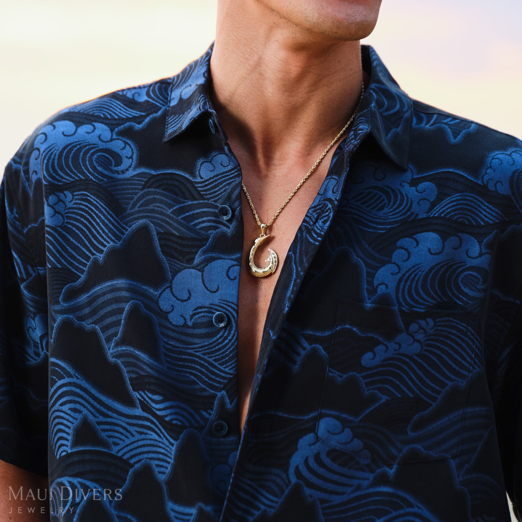 Man wearing a blue patterned shirt with a Hawaiian fish hook pendant in gold
