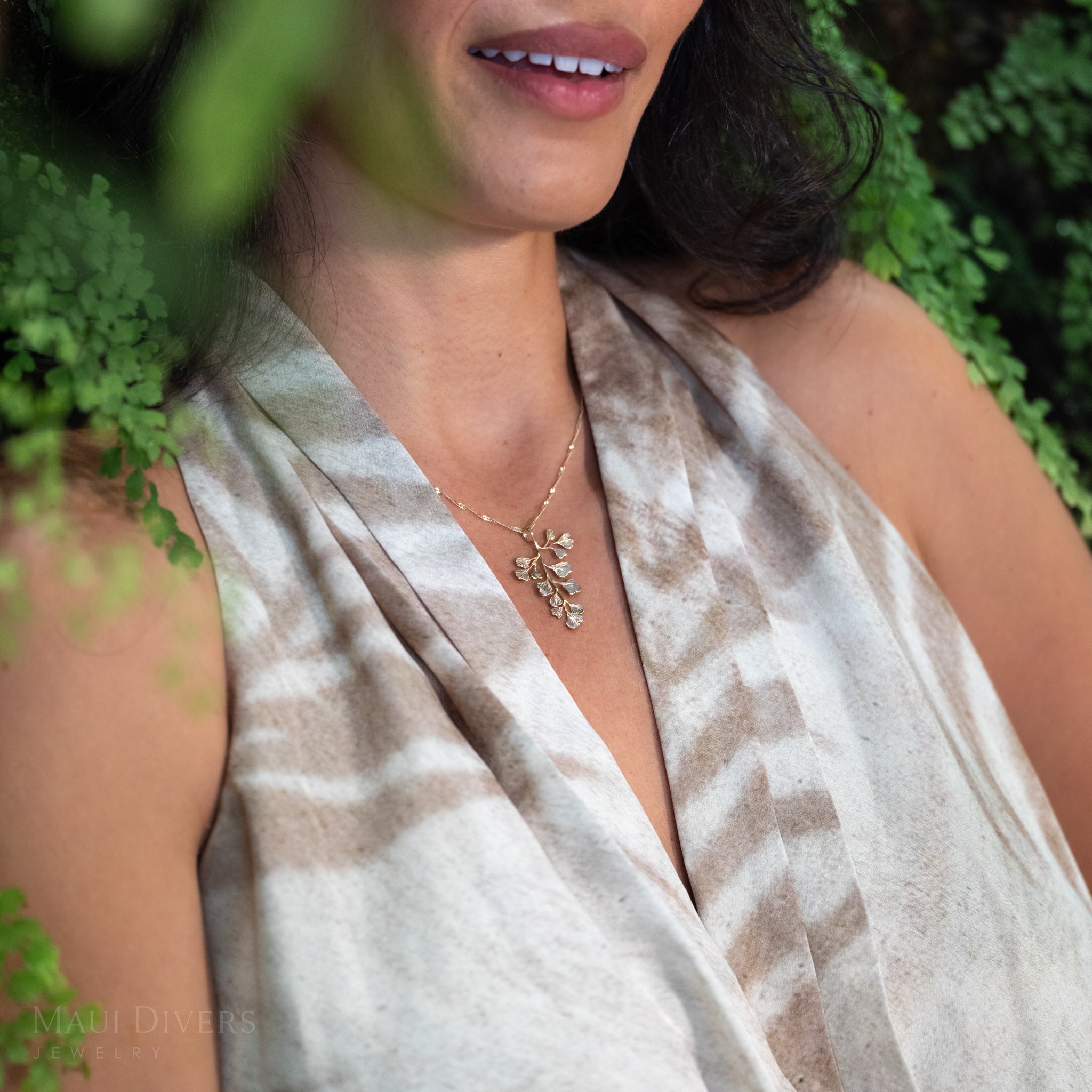 A close-up lifestyle shot of a woman wearing a delicate 14k yellow gold maiden hair fern pendant on a Singapore chain over a beige-and-brown patterned halter top, set against a soft-focus background of maiden hair plants.