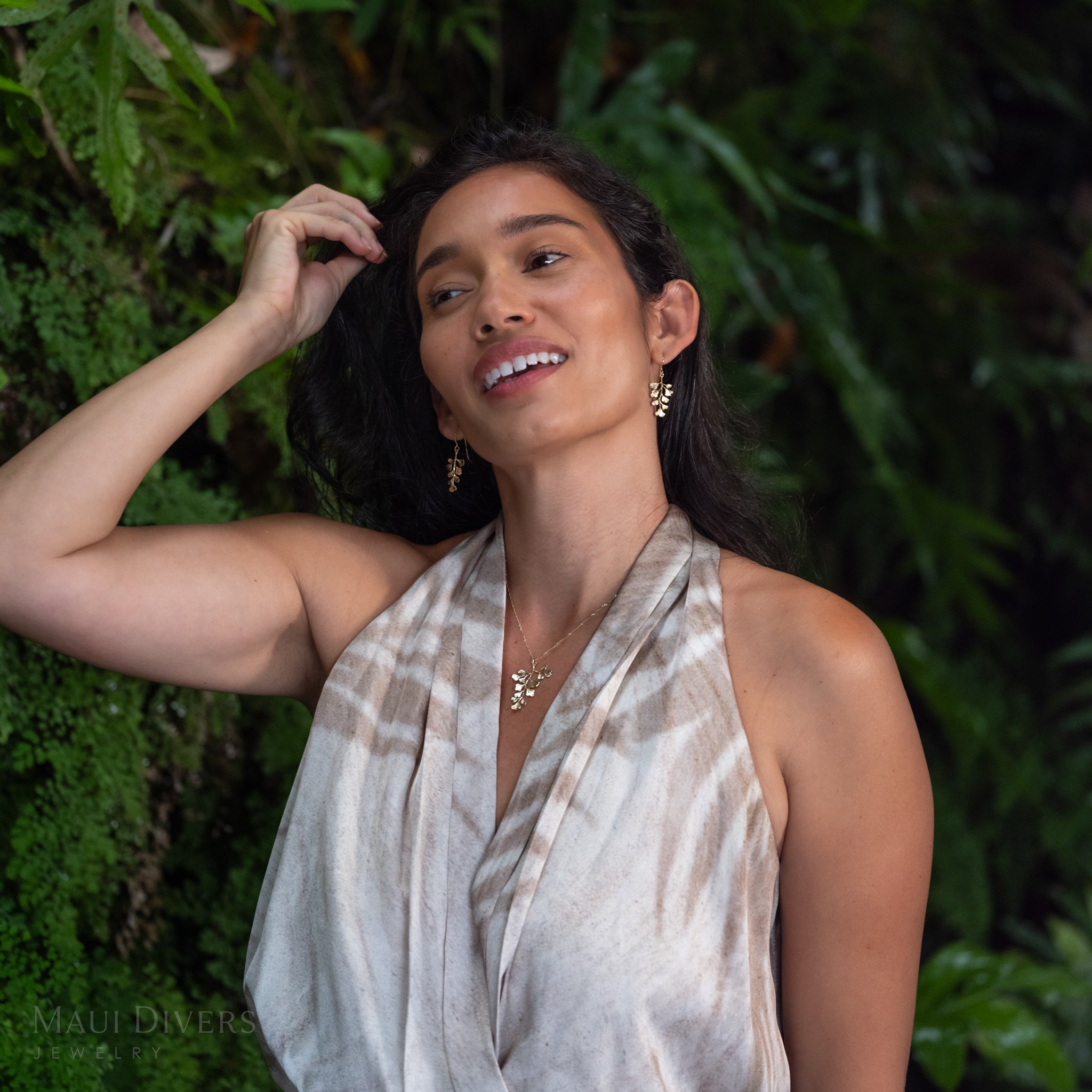 A woman wearing a maiden hair fern pendant in 14k yellow gold on a singapore chain and matching earrings, beige and white patterned halter wrap top, posing outdoors against a lush green foliage background.