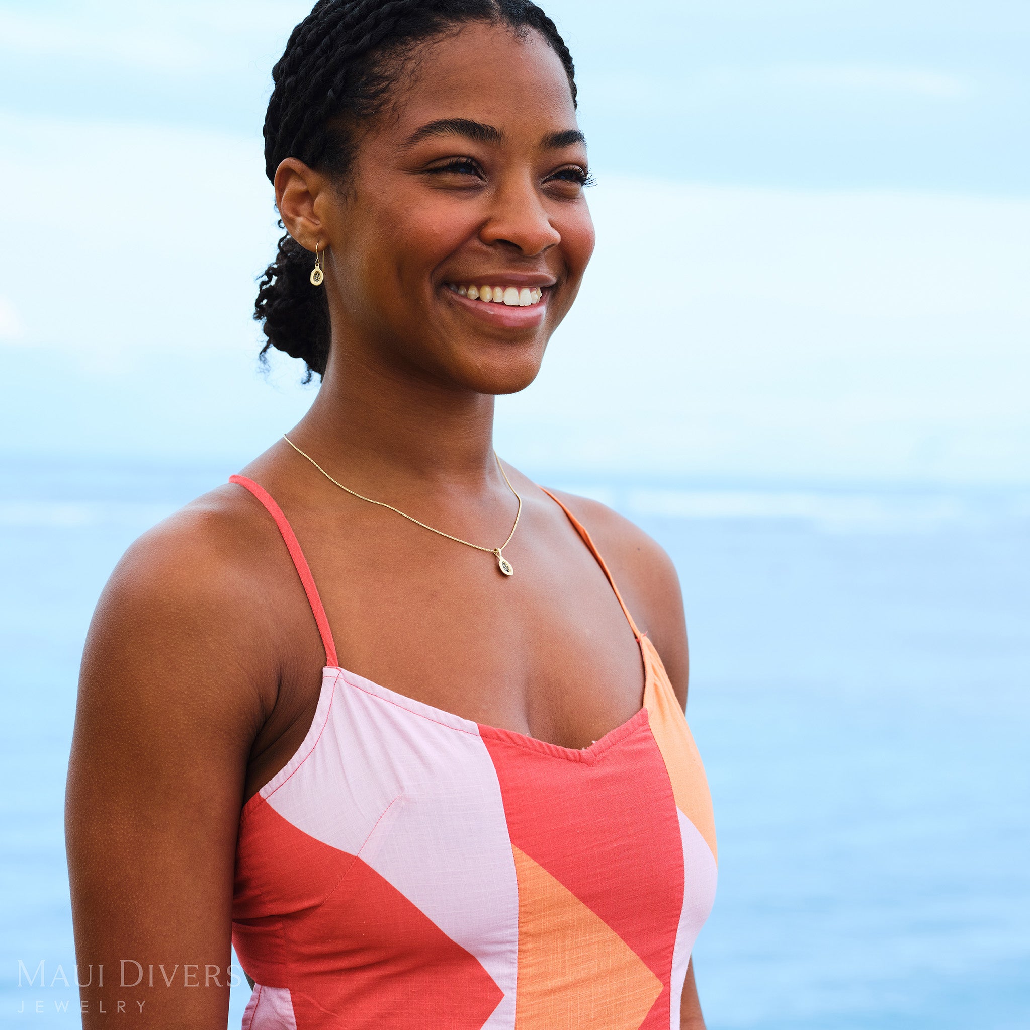 Smiling woman in an orange and pink top wearing a Cute Fruits Papaya Pendant in 14k yellow gold with black diamonds around her neck and matching Cute Fruits Papaya Earrings in 14k yellow gold with black diamonds, against a blurred ocean background
