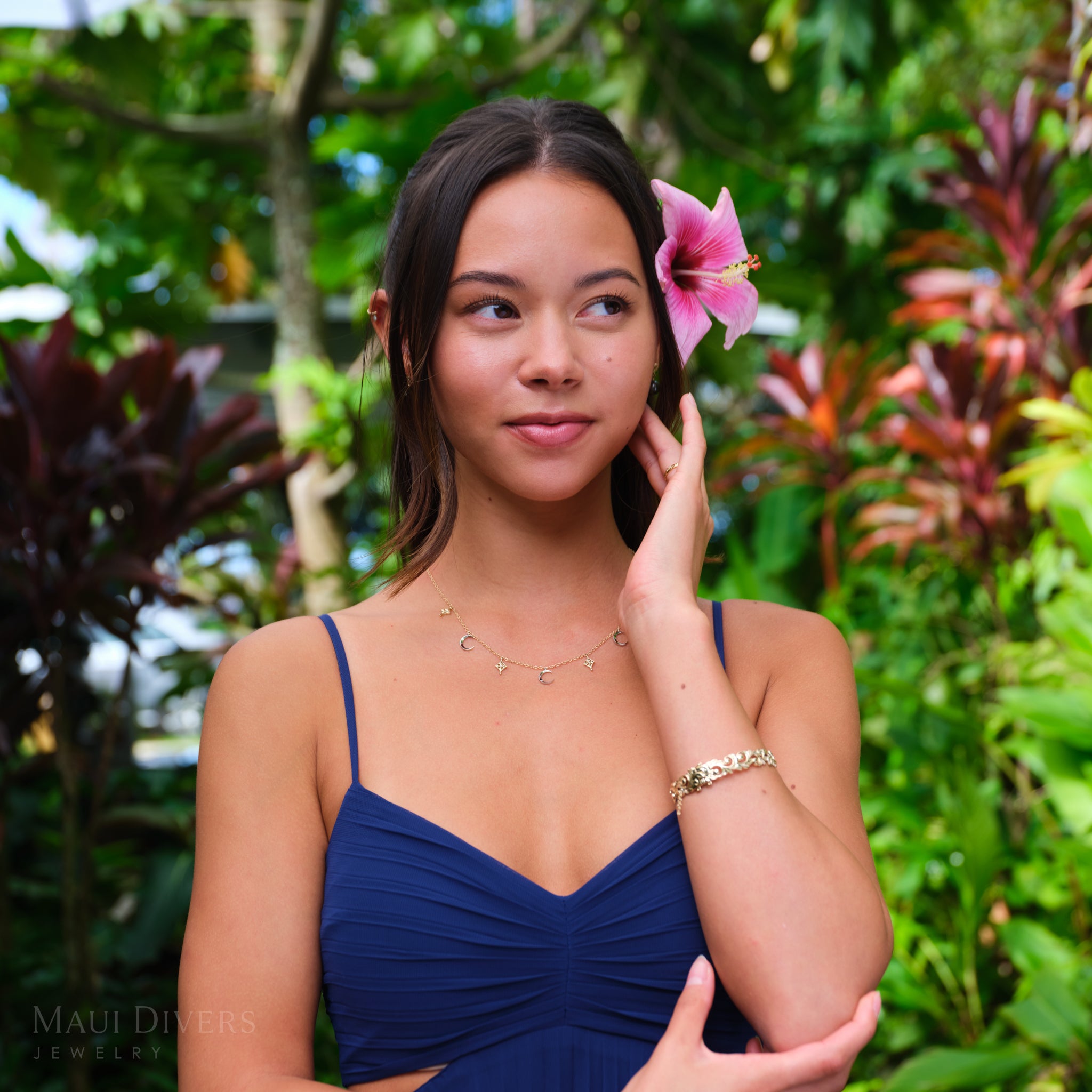 Woman in a blue dress and hibiscus flower in her ear, wearing moon and star necklace in white and yellow gold