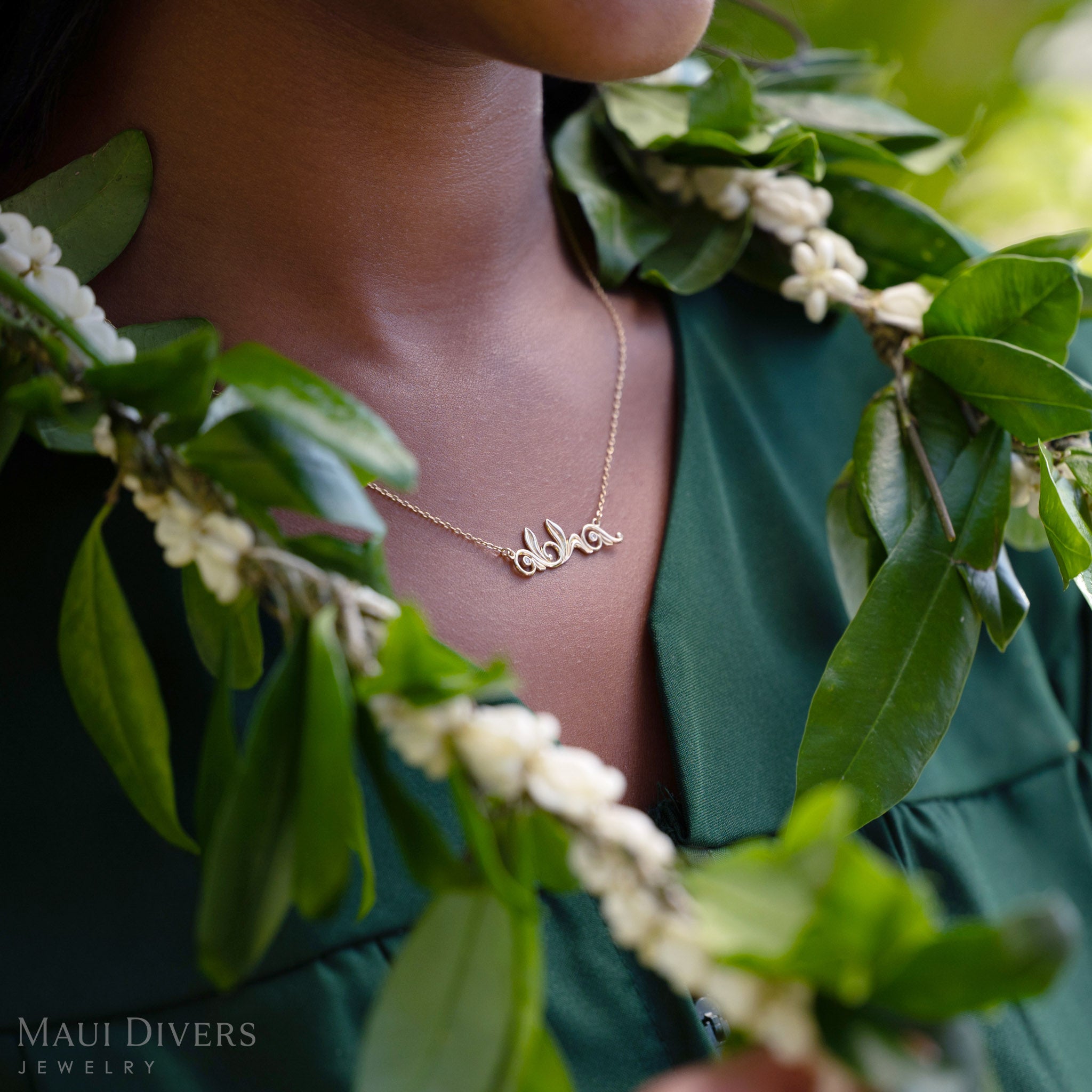 Close-up of a person in a green top wearing a Living Heirloom Aloha necklace in 14k yellow gold with diamond accents, with a green lei intertwined with white crown flowers