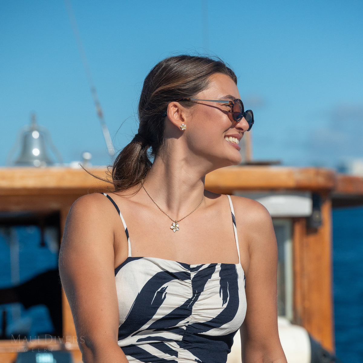 A woman in a patterned navy and white top wearing a gold plumeria pendant with mother of pearl inlay on an ovalina chain, with a wooden boat and ocean in the background