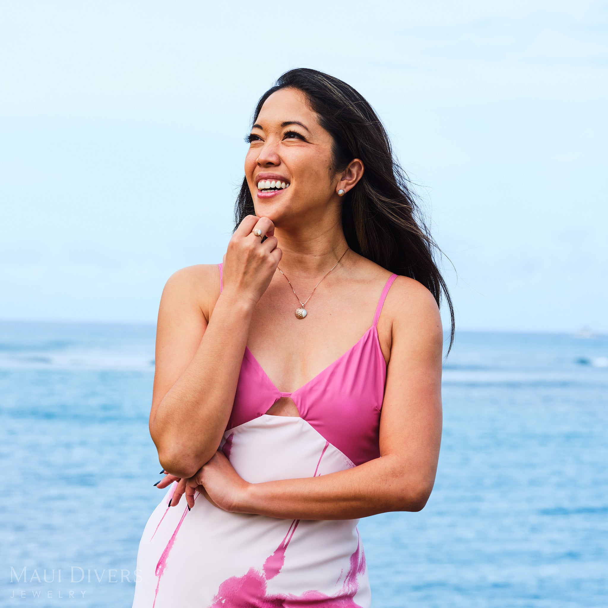 Smiling woman in a pink dress wearing a matching set of Cute Fruits Lychee Mother of Pearl earrings, ring, and pendant in 14k rose gold against a blurred ocean background