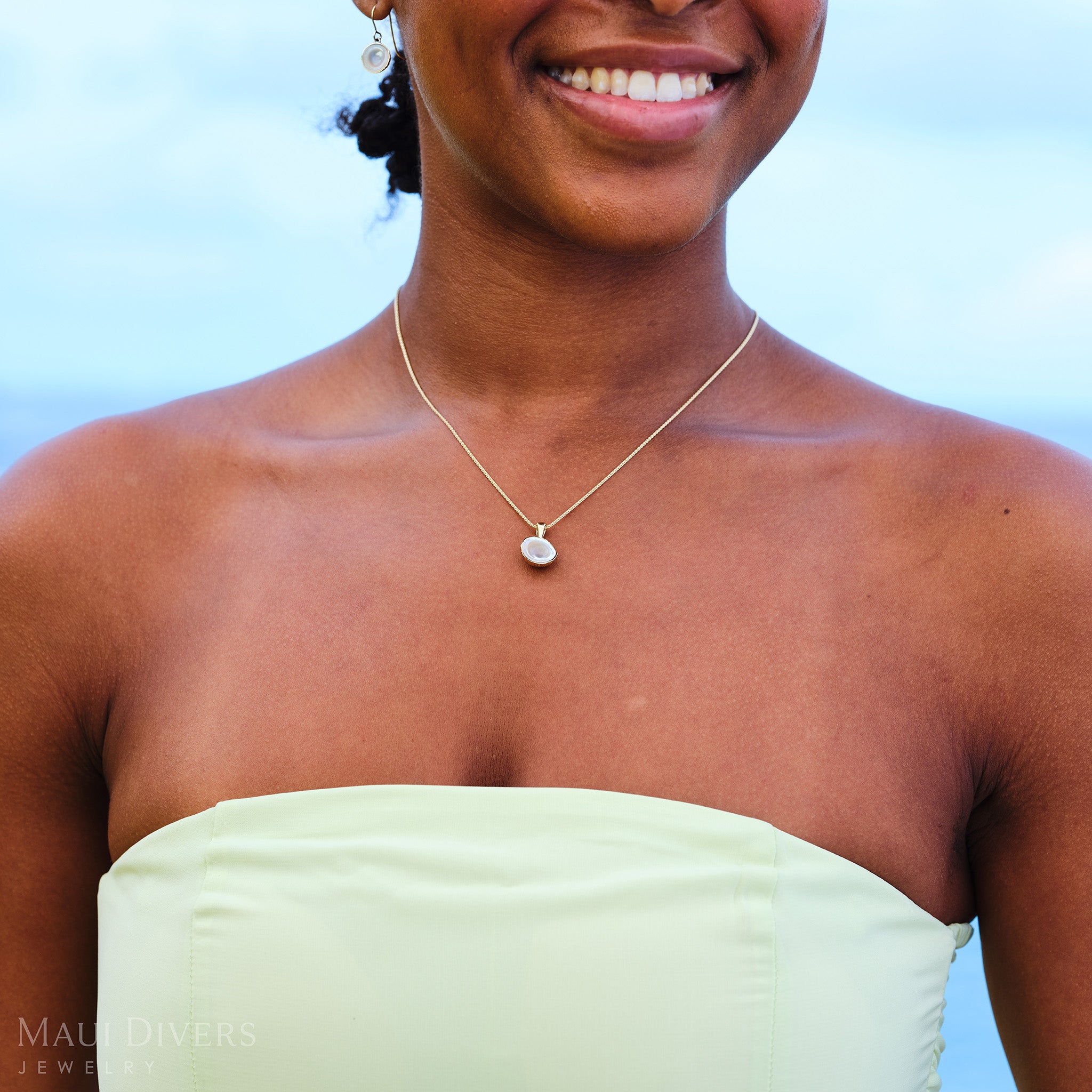 Close-up of a smiling woman in a pastel green dress wearing a Cute Fruits Coconut Mother of Pearl Pendant in 14k yellow gold