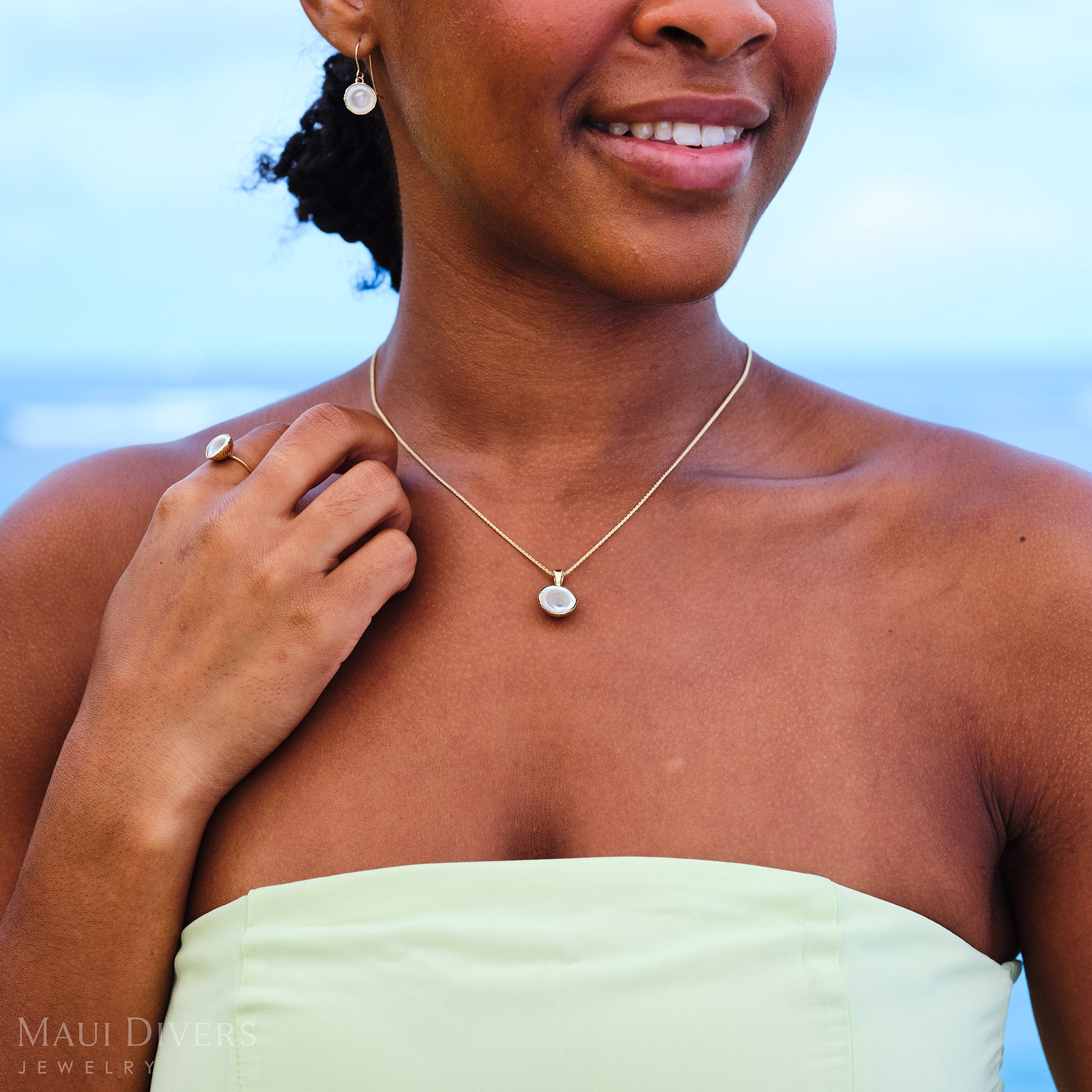 Close-up of a smiling woman in a pastel green dress wearing a Cute Fruits Coconut Mother of Pearl matching set with a pendant, ring, and earrings, all in 14k yellow gold