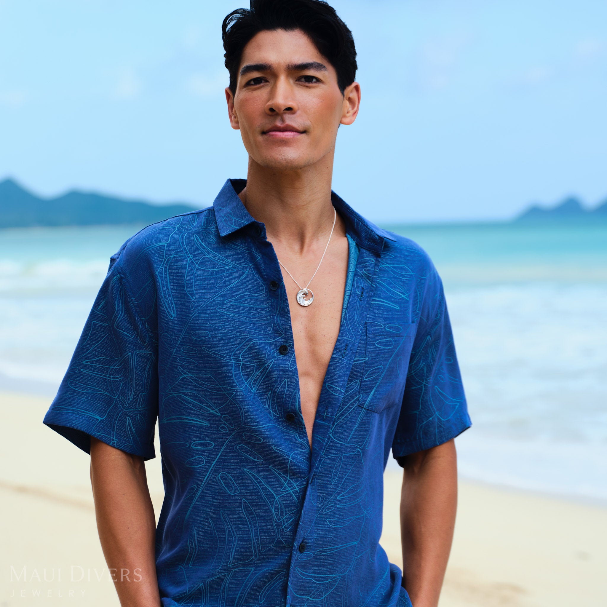 Man wearing a silver wave necklace and blue patterned shirt on a beach with ocean and sky in the background