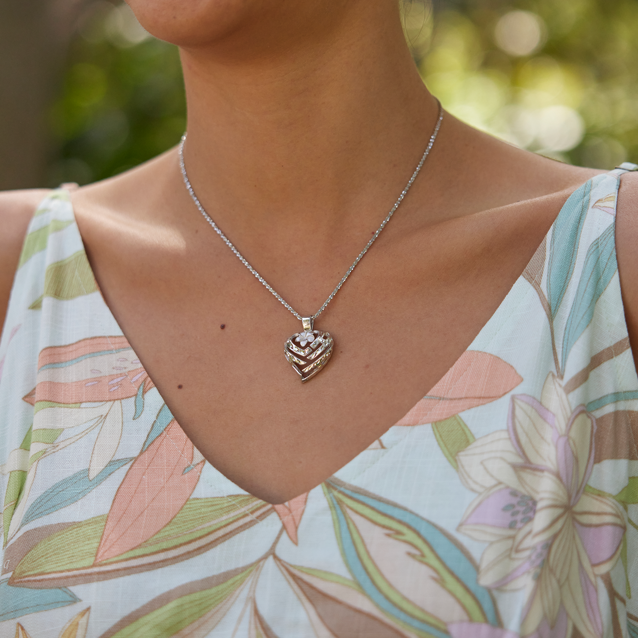 Close up of an Aloha Heart Plumeria Pendant in Sterling Silver on a woman's chest with blurred trees in the back