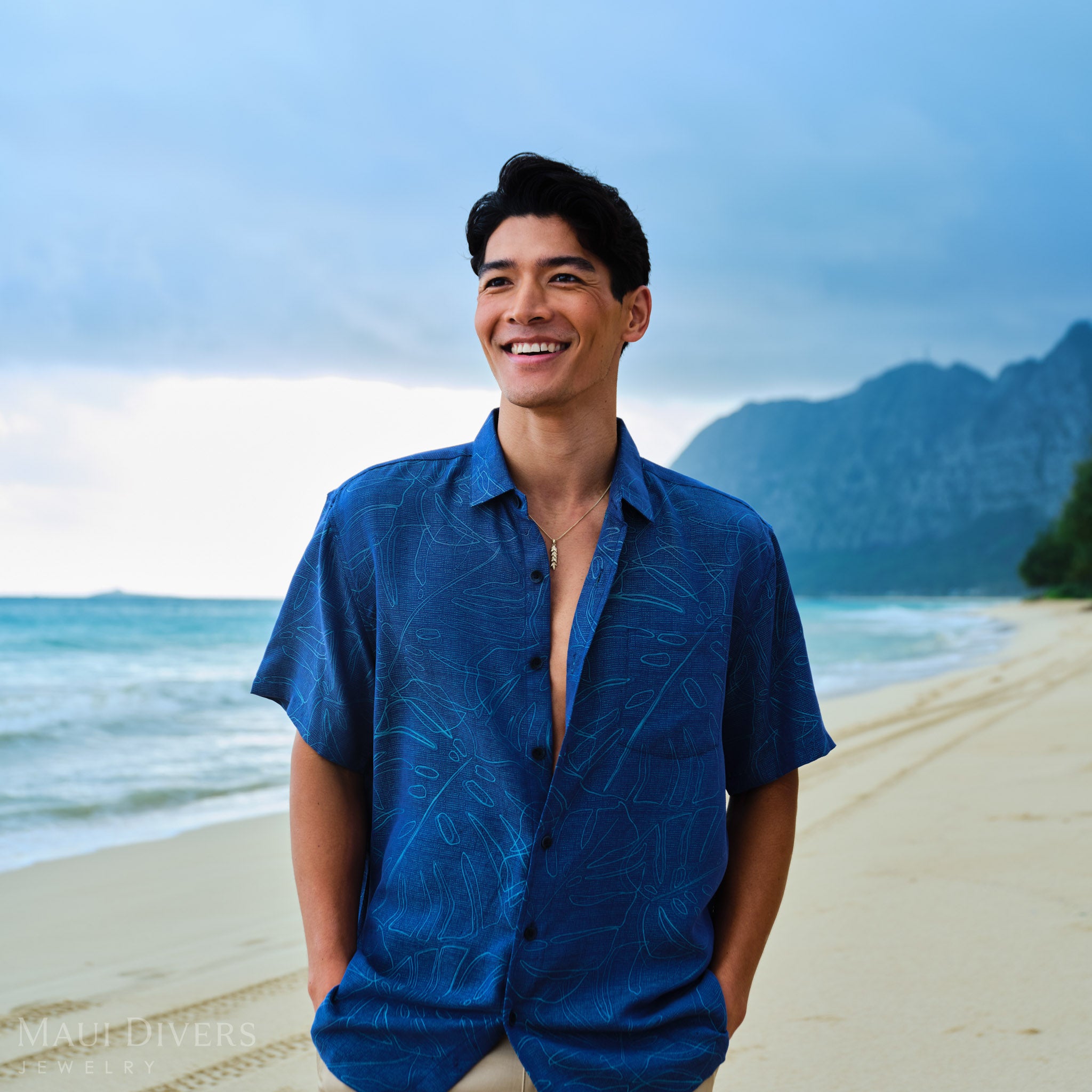 Man wearing a blue patterned shirt with a gold maile leaf pendant on a chain on a beach with mountains in the background